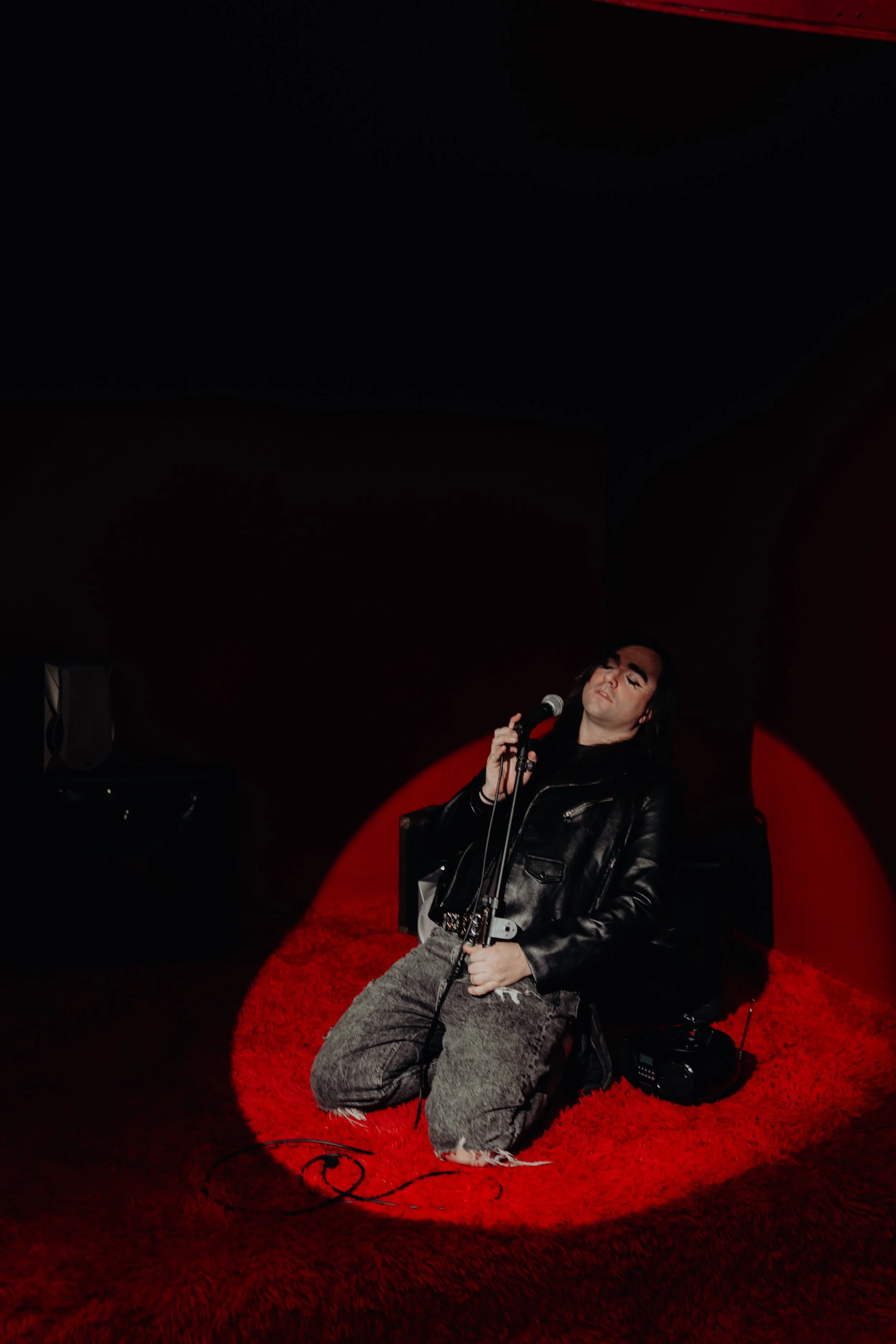 A person kneeling on a red shaggy rug in a dimly lit stage or performance space, holding a microphone and music stand, dressed in a leather jacket and gray pants with a camo pattern, with a black speaker and musical equipment nearby.