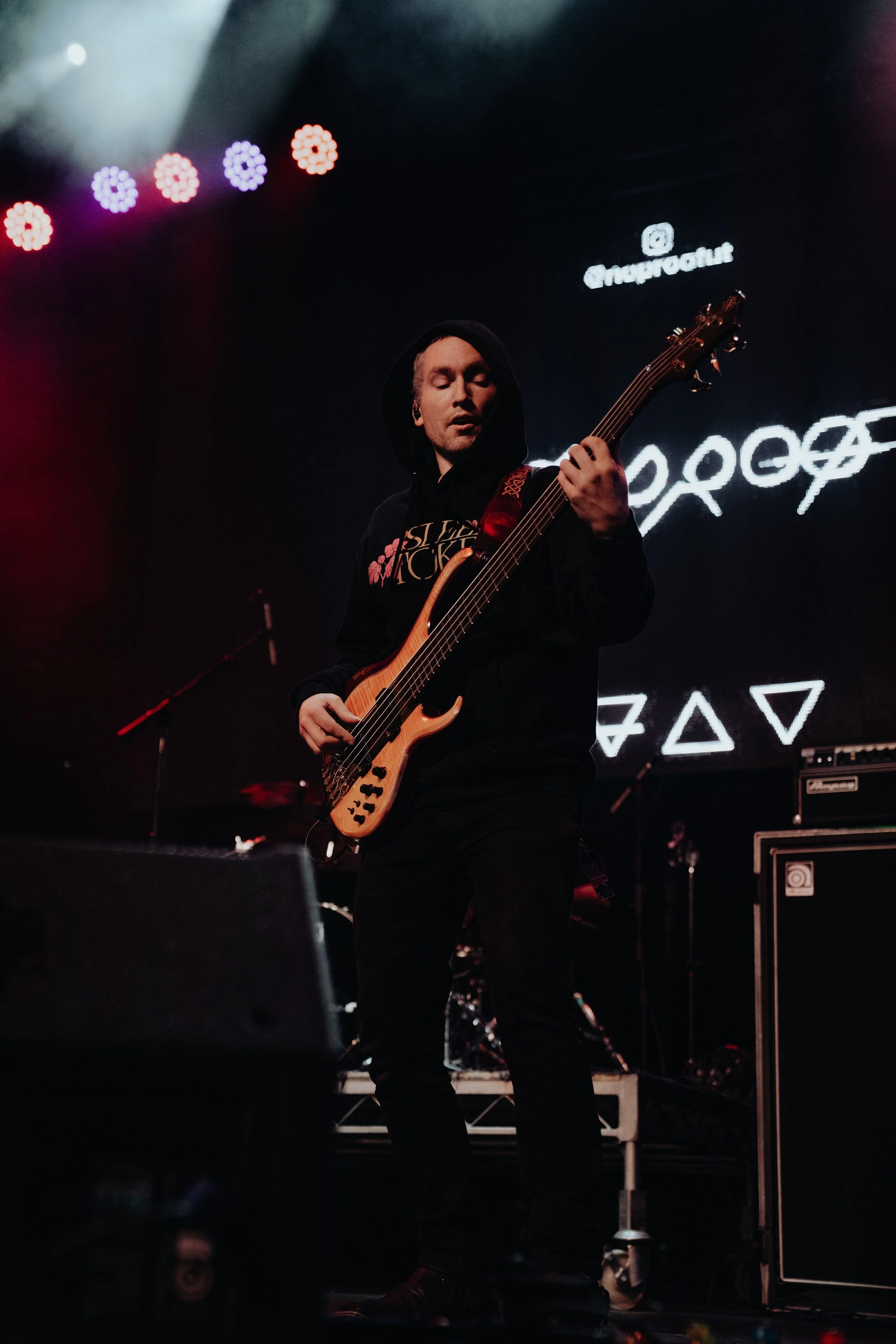 A person playing an electric bass guitar on stage with colorful lights and a digital screen in the background showing shapes and symbols.