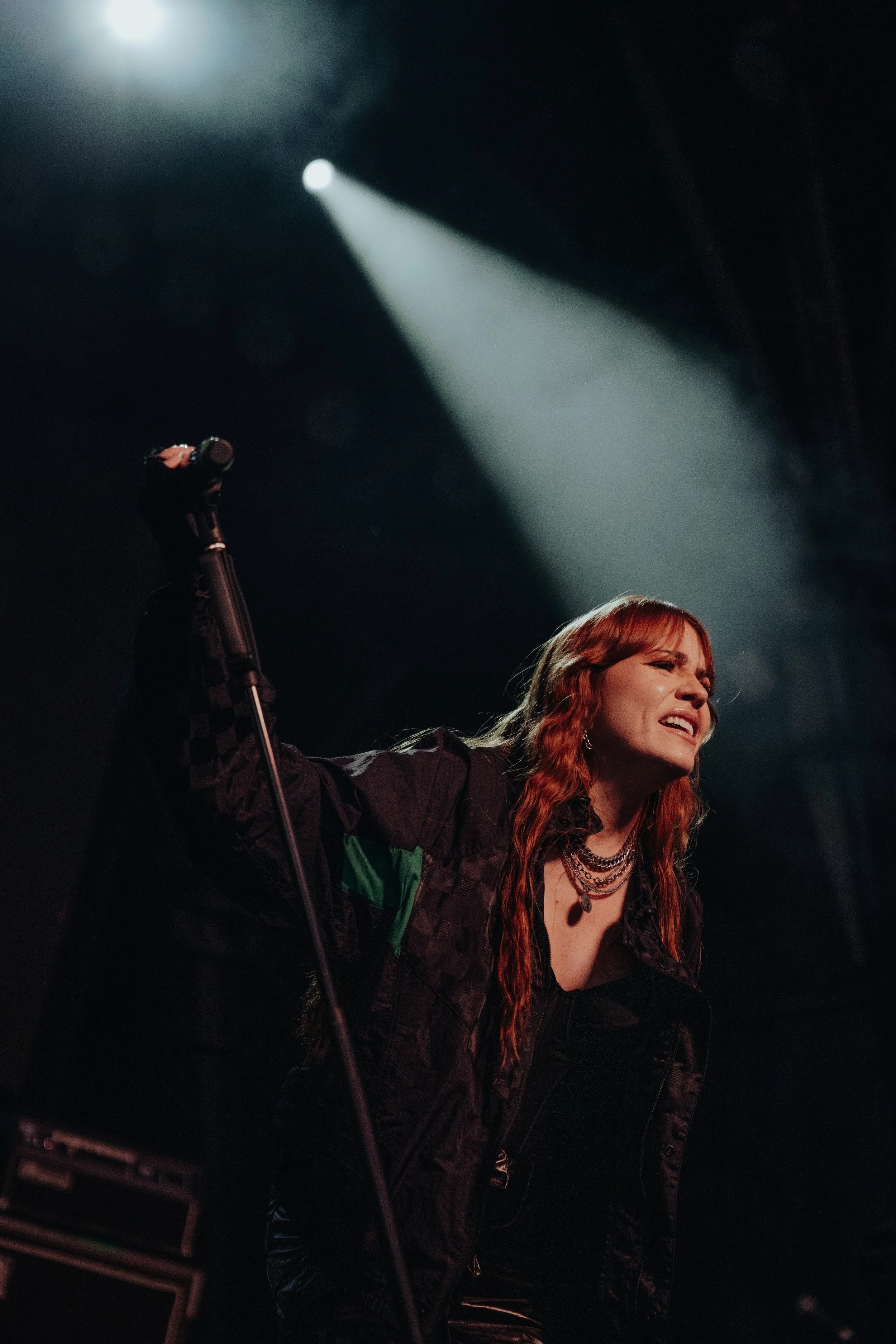 A woman with long red hair singing into a microphone, wearing layered necklaces and a black jacket with green and black checkered sleeves, on stage with dramatic spotlighting.