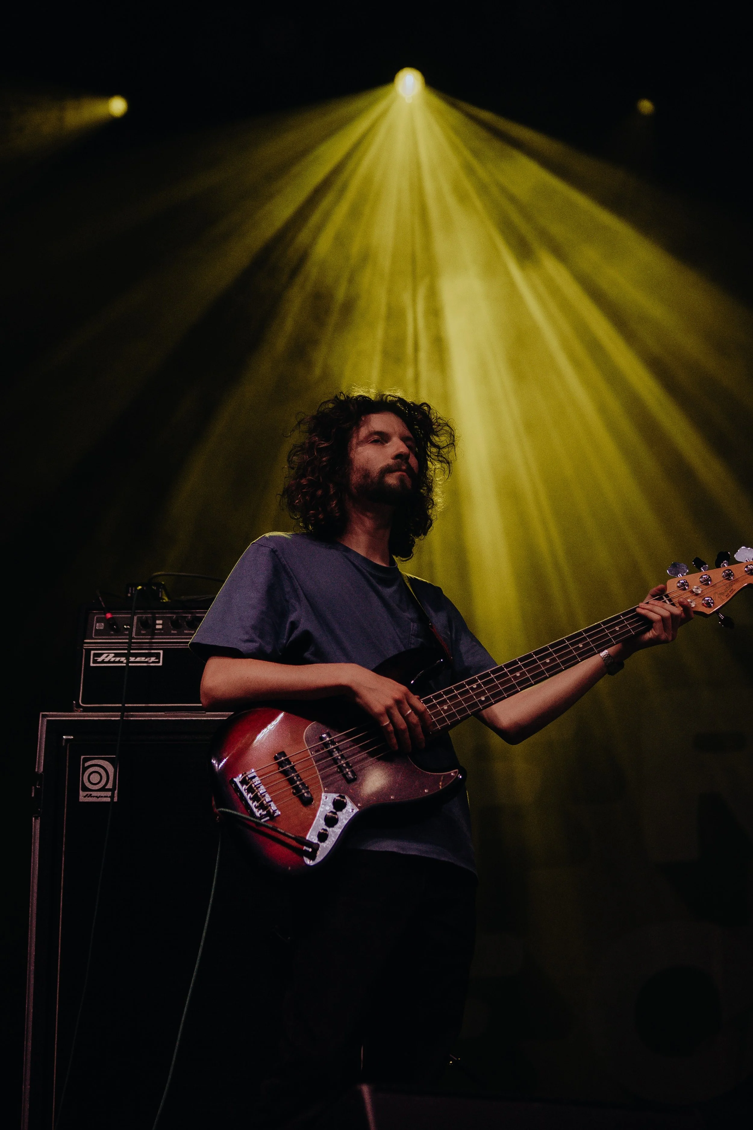 A man with curly hair and a beard playing an electric bass guitar on a stage, with yellow stage lights and audio equipment in the background.