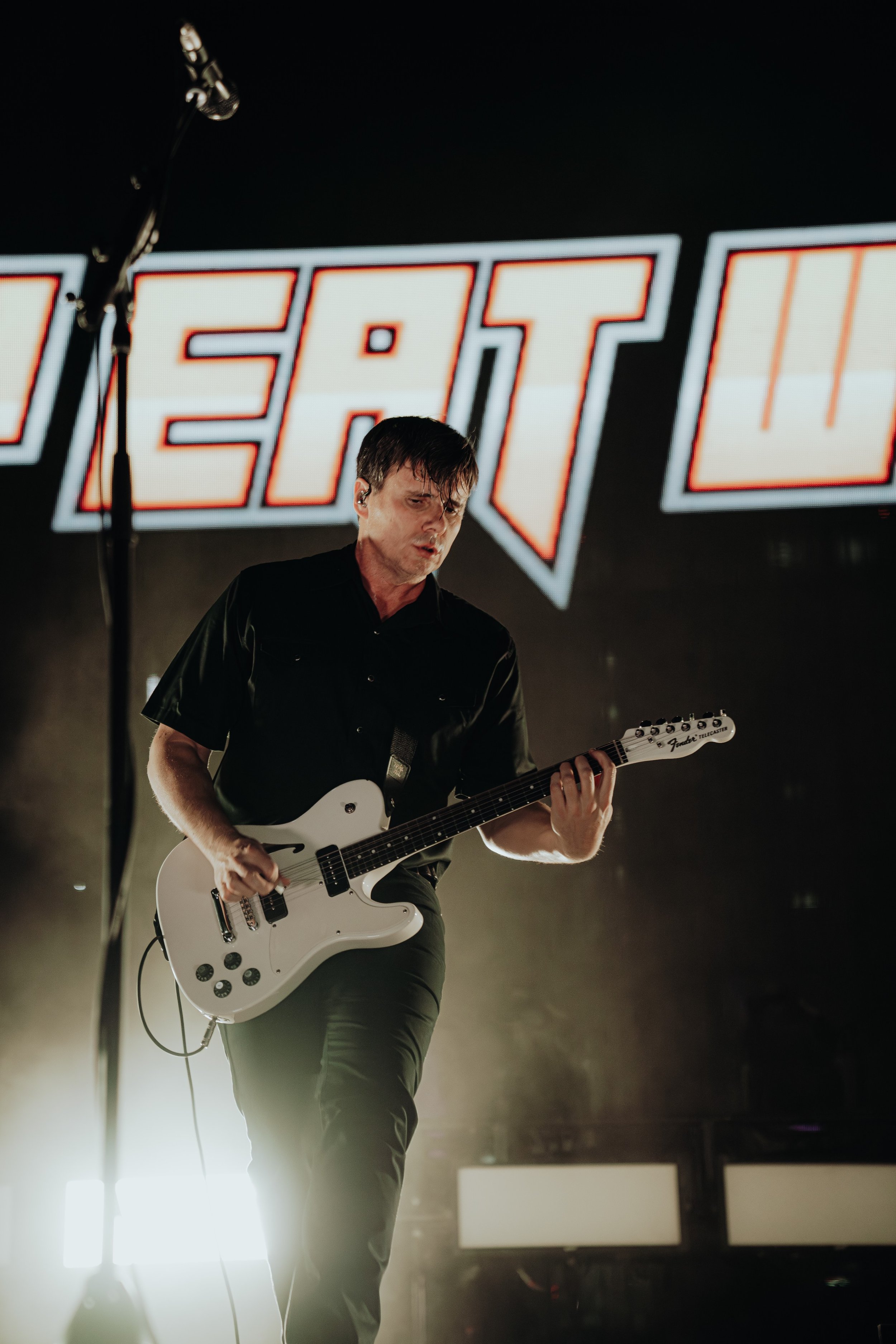 A male guitarist playing electric guitar on stage with large illuminated text in the background and stage lights shining from behind him.
