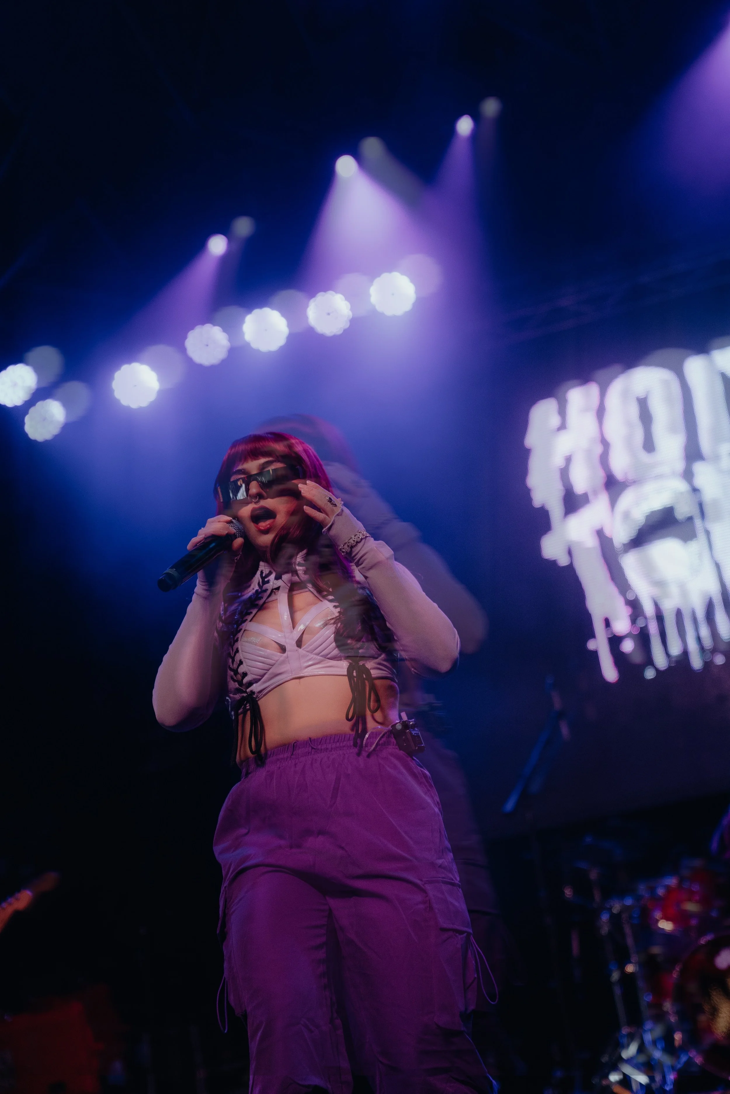 Performer on stage with purple lighting, wearing dark sunglasses, purple cargo pants, beige top with black lace-up detail, singing into a microphone.