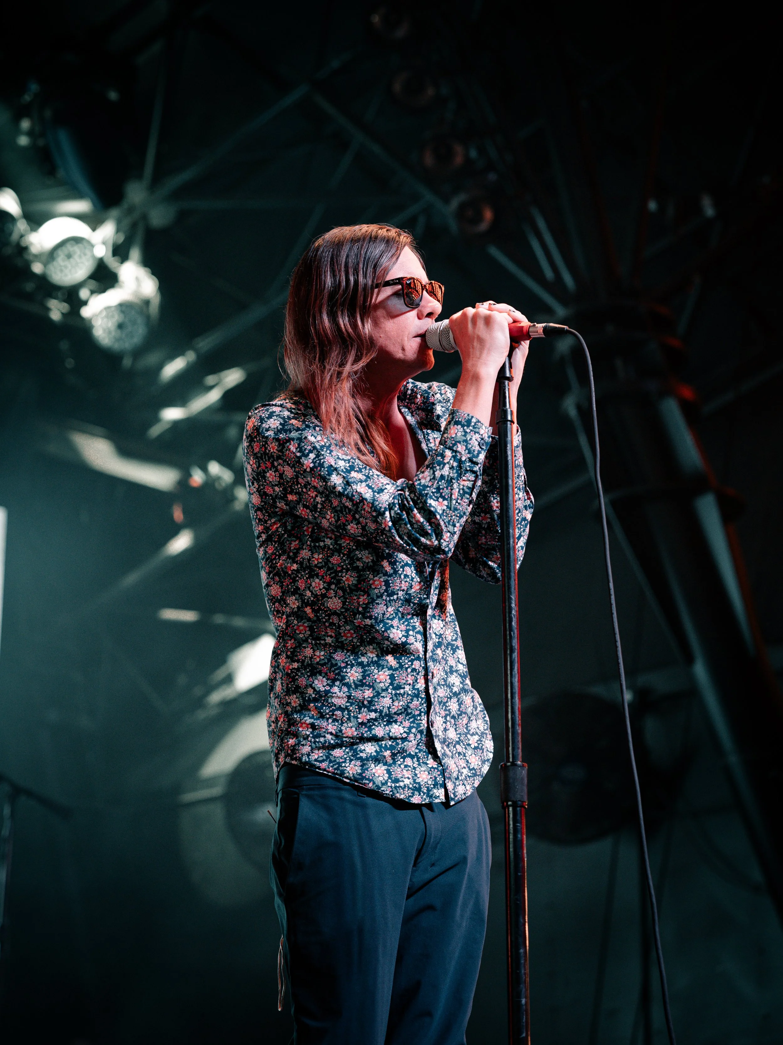 A woman singing into a microphone on stage, wearing sunglasses, a floral shirt, and dark pants, with stage lights and equipment in the background.
