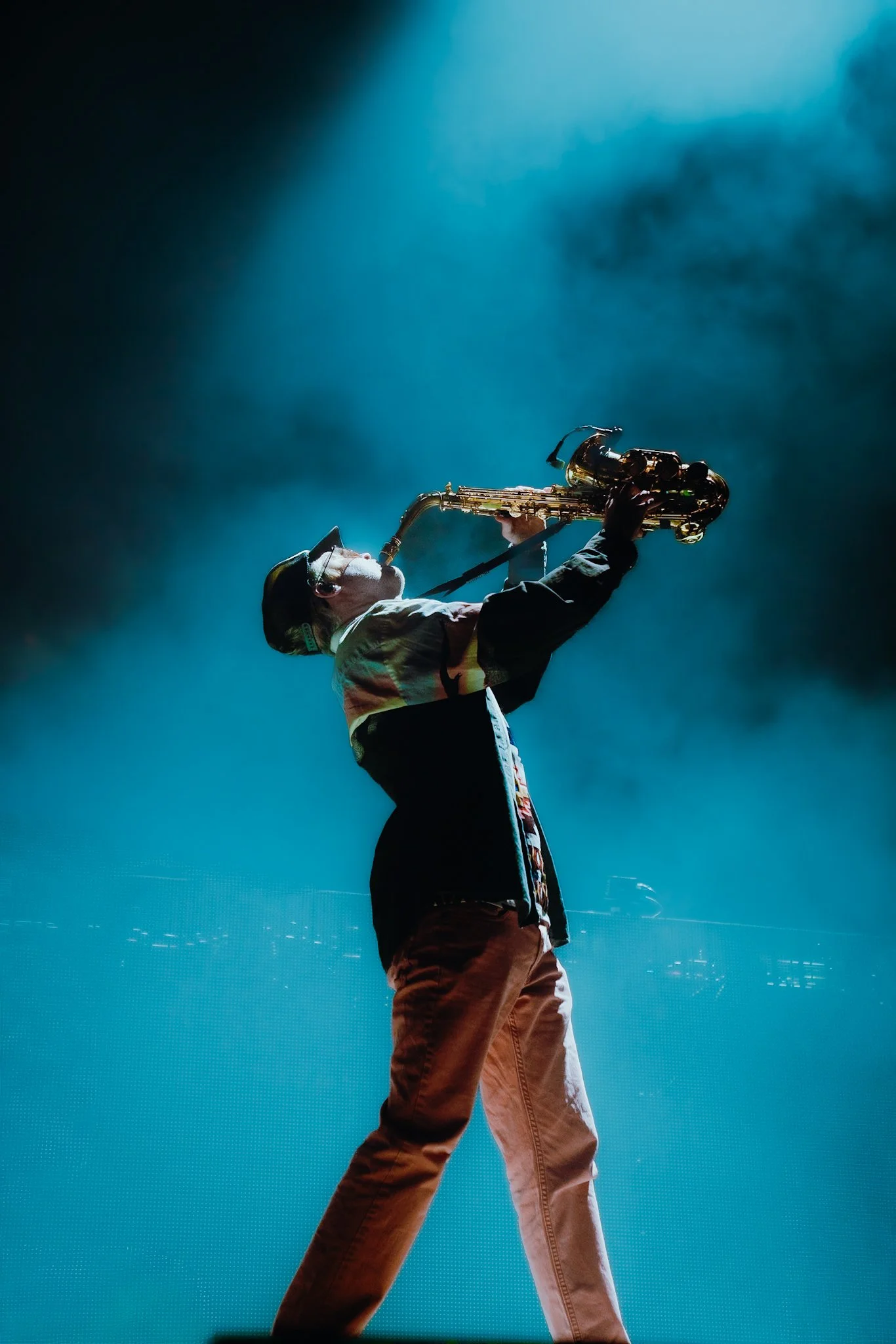 A person wearing a baseball cap and glasses playing a saxophone on stage, with a dark background and blue lighting.