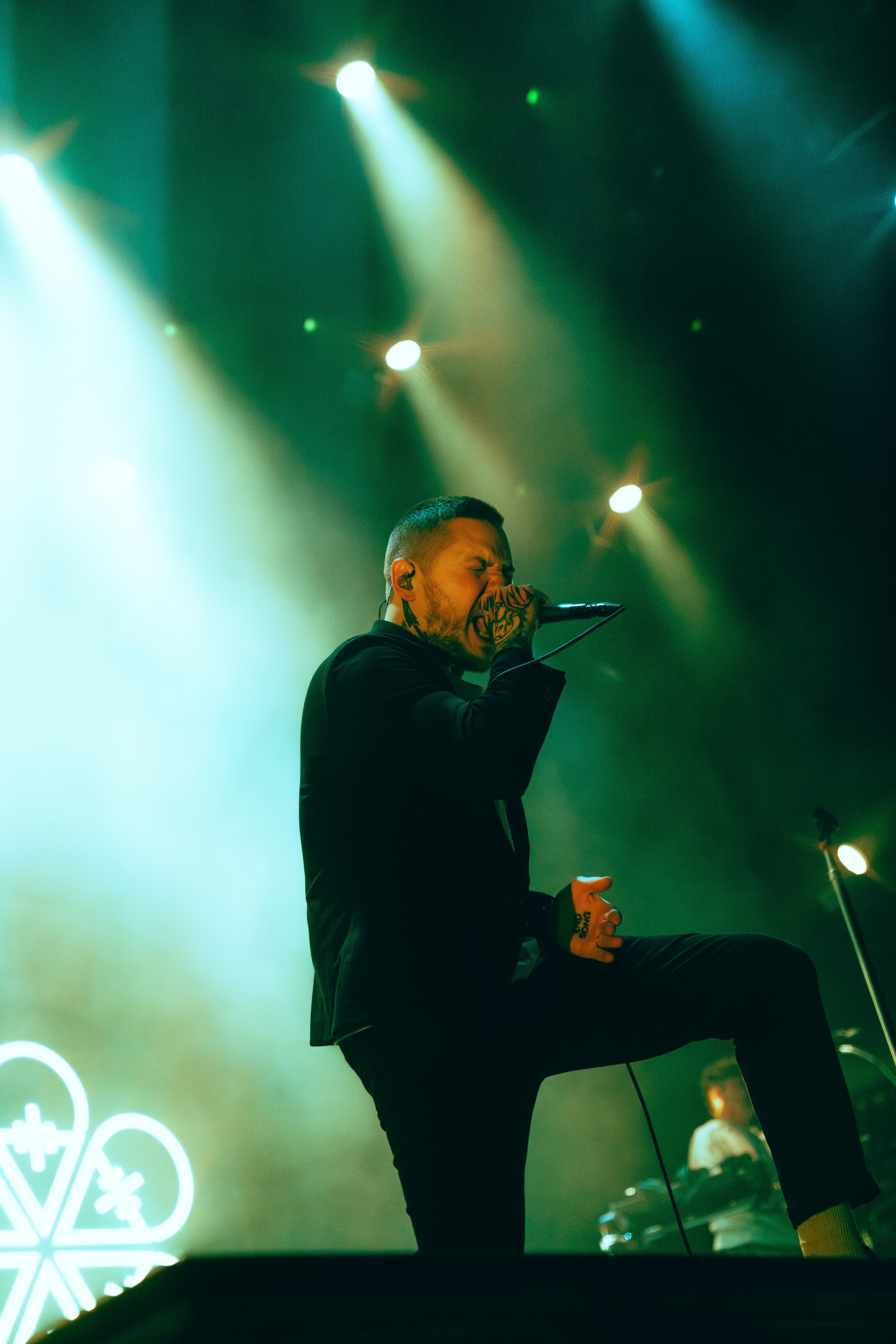 A man with tattoos singing into a microphone on stage with green and yellow stage lights, wearing a black jacket and pants, with a neon sign featuring a snowflake shape in the background.