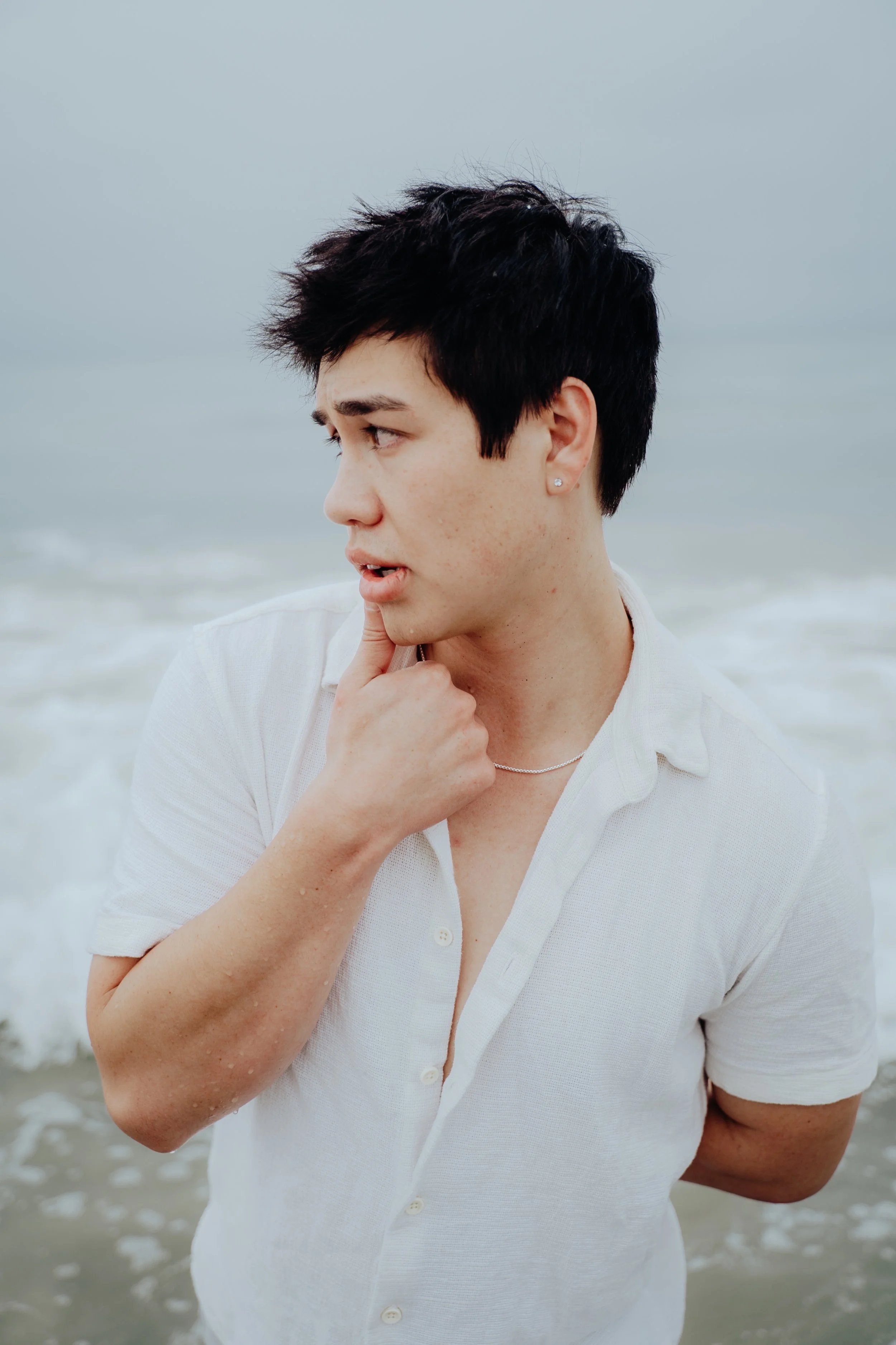 A young person with short dark hair and earrings, wearing a white shirt, standing in the ocean near the shore, looking to the left with their hand resting on their chin.