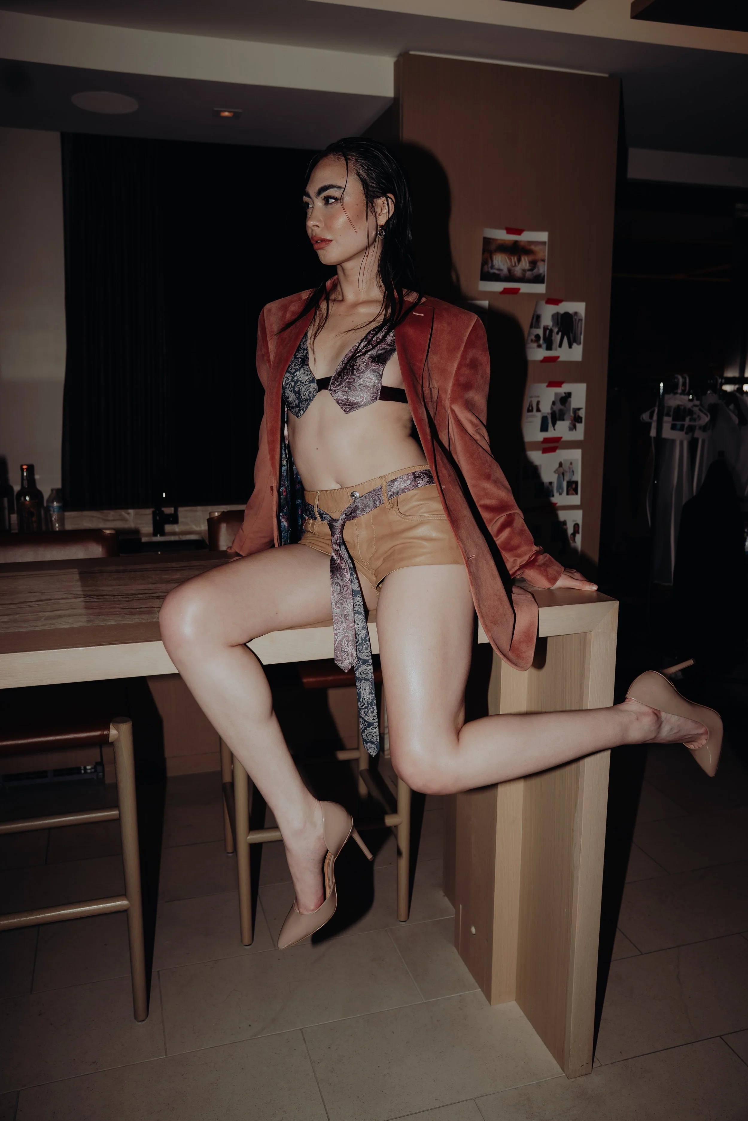 A woman in business attire, partially undressed, leaning against a table in a dimly lit room, with her legs raised on a chair, and photos taped on the wall in the background.