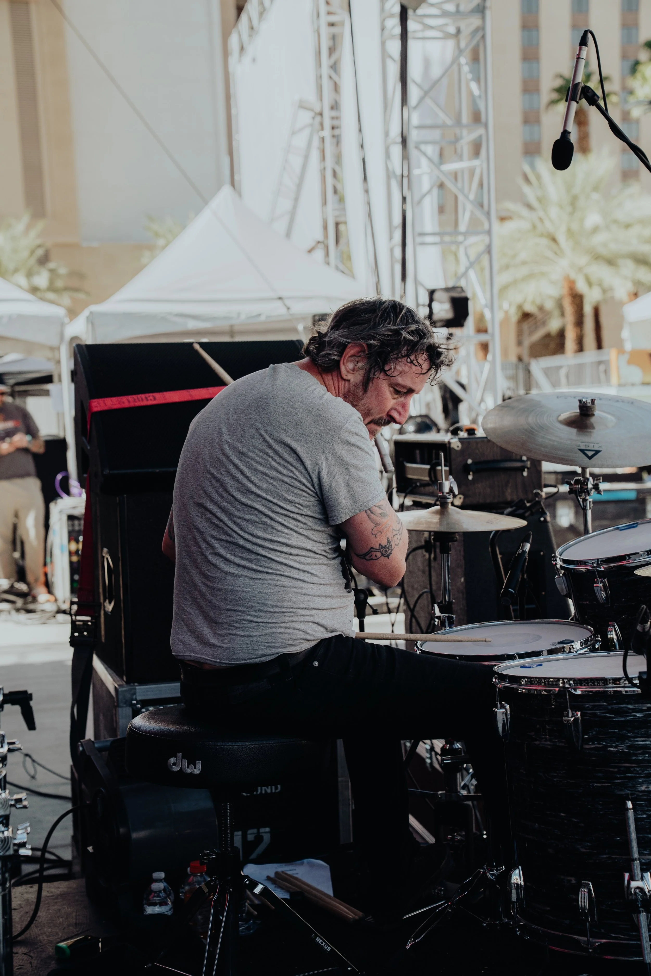 Musician playing drums on an outdoor stage surrounded by equipment and setup, with tents and buildings in the background.