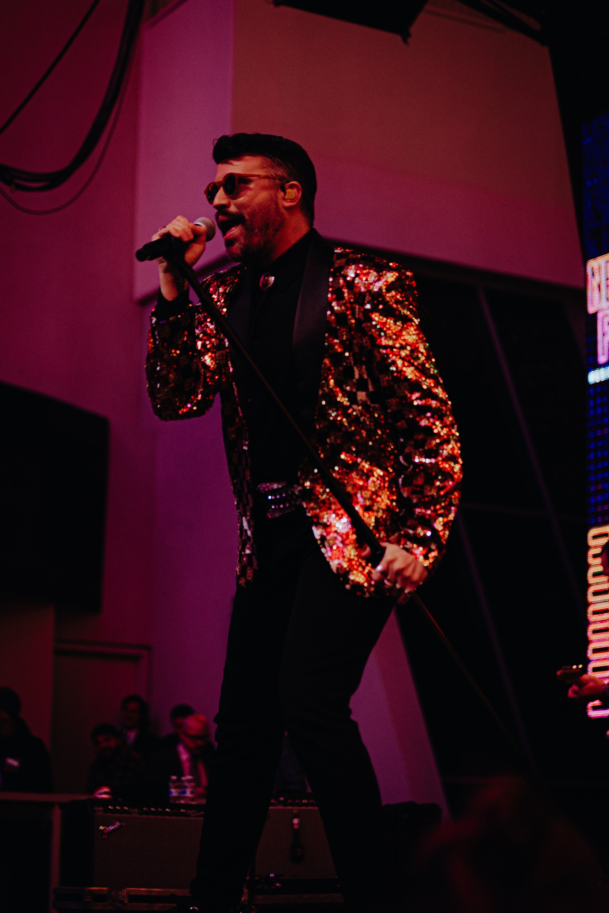 A male performer singing into a microphone during a concert, wearing black sunglasses and a sparkly, sequined blazer with a checkered tie, on a stage with pink and purple lighting.