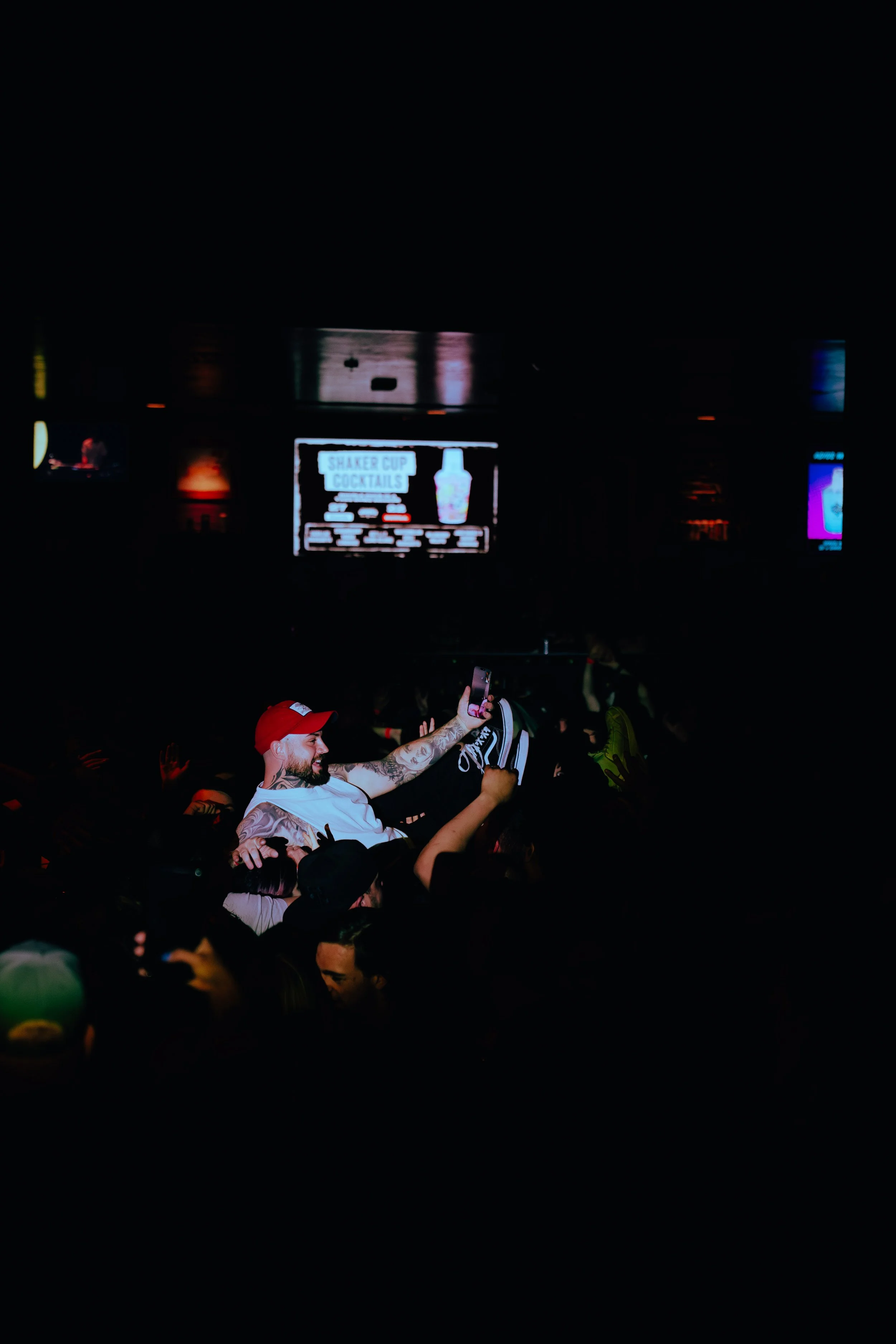 Crowd at a nightclub with a person on shoulders taking a selfie, multiple screens and signs visible, including a sign for shaken cup cocktails.