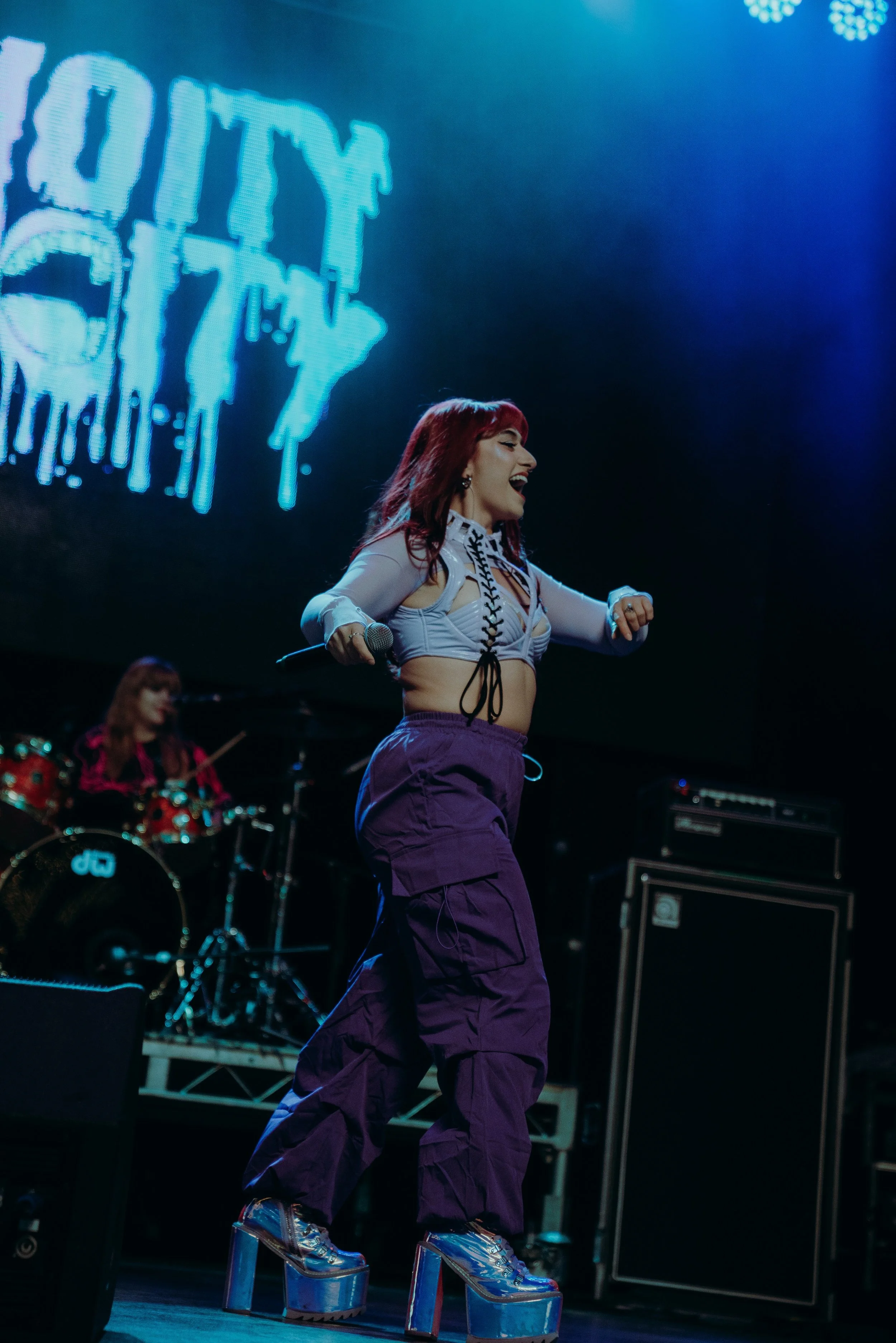 A female performer with red hair singing into a microphone on stage, wearing a white top and purple cargo pants, standing on tall platform shoes. In the background, a drummer playing a drum kit and a large LED screen displaying blue graphics are visi