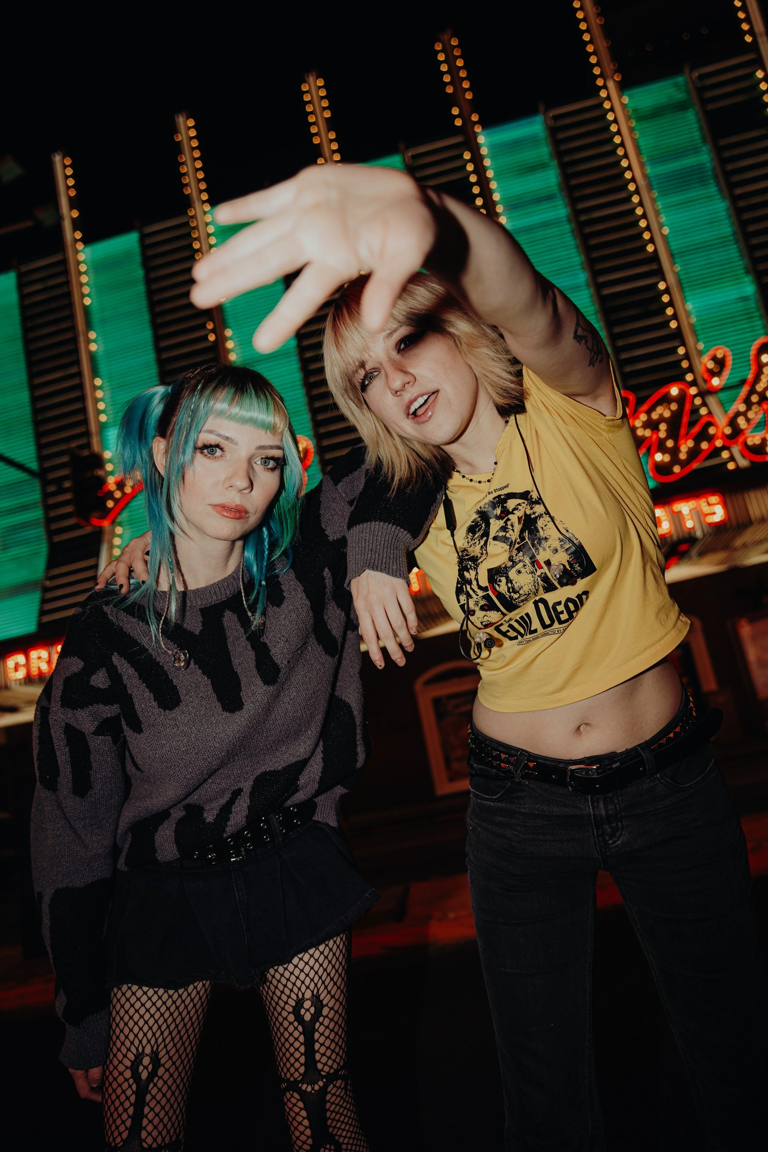 Two women in a nightclub, one with colorful hair and dark makeup, wearing a animal print sweater, fishnet tights, and a black skirt; the other with blonde hair and a yellow T-shirt, posing with her arm extended. Neon lights and a sign are visible in 