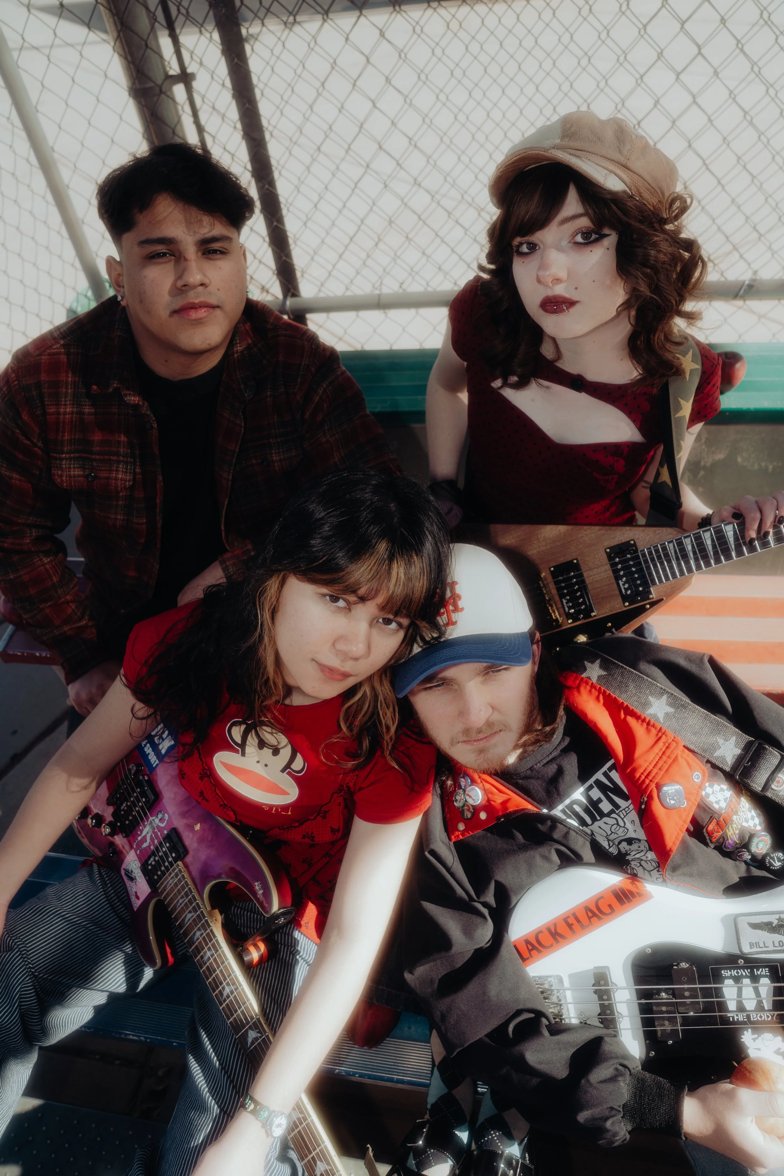 Group of five young people sitting on a bench, some holding guitars, at an outdoor location with chain-link fencing in the background.