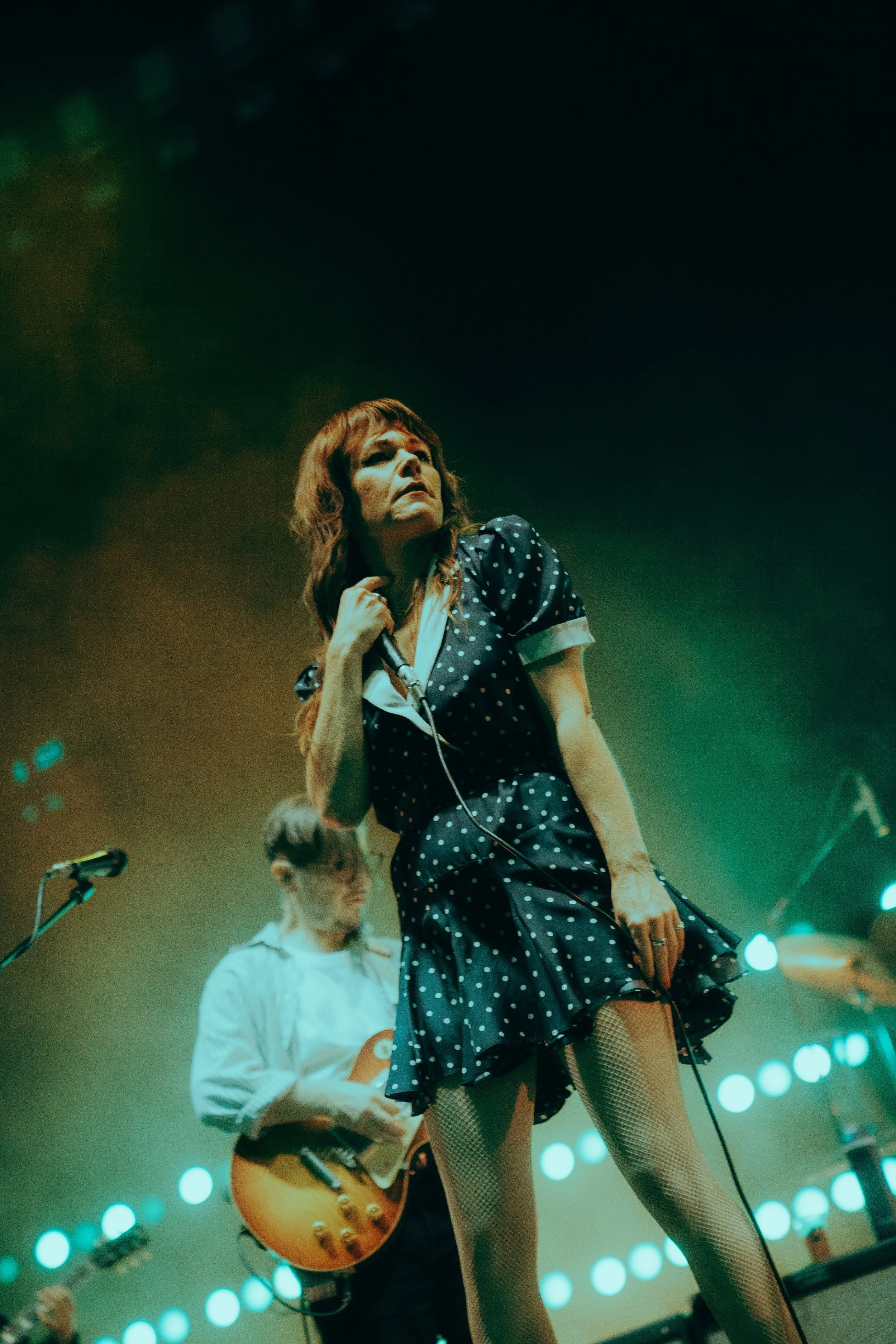 A woman singing into a microphone during a concert, dressed in a polka dot dress and fishnet stockings, with a guitarist playing in the background against a backdrop of green and blue stage lights.
