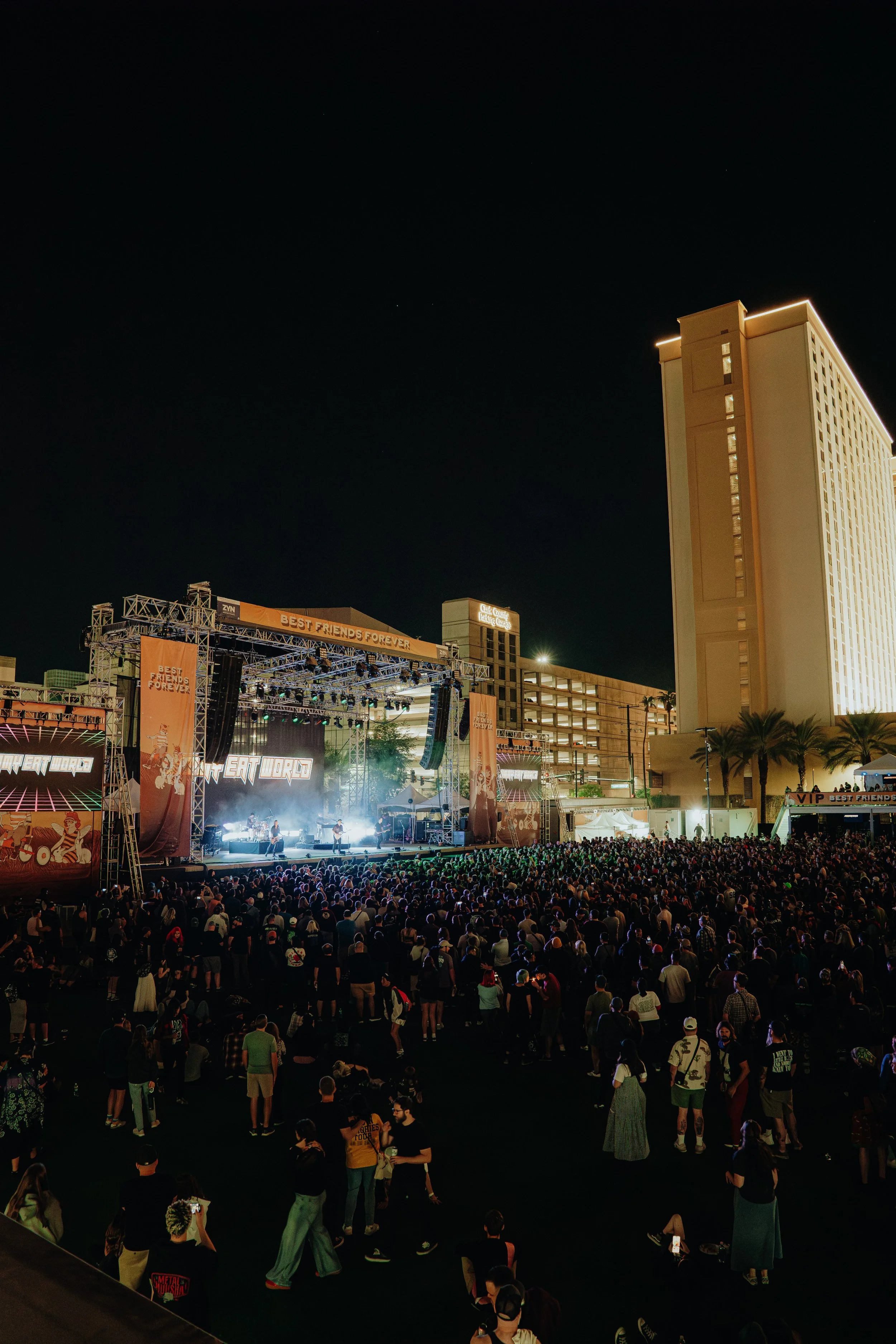 Nighttime outdoor concert in a city with a large crowd, stage with performers, tall hotel building with lights, and several smaller buildings and palm trees.