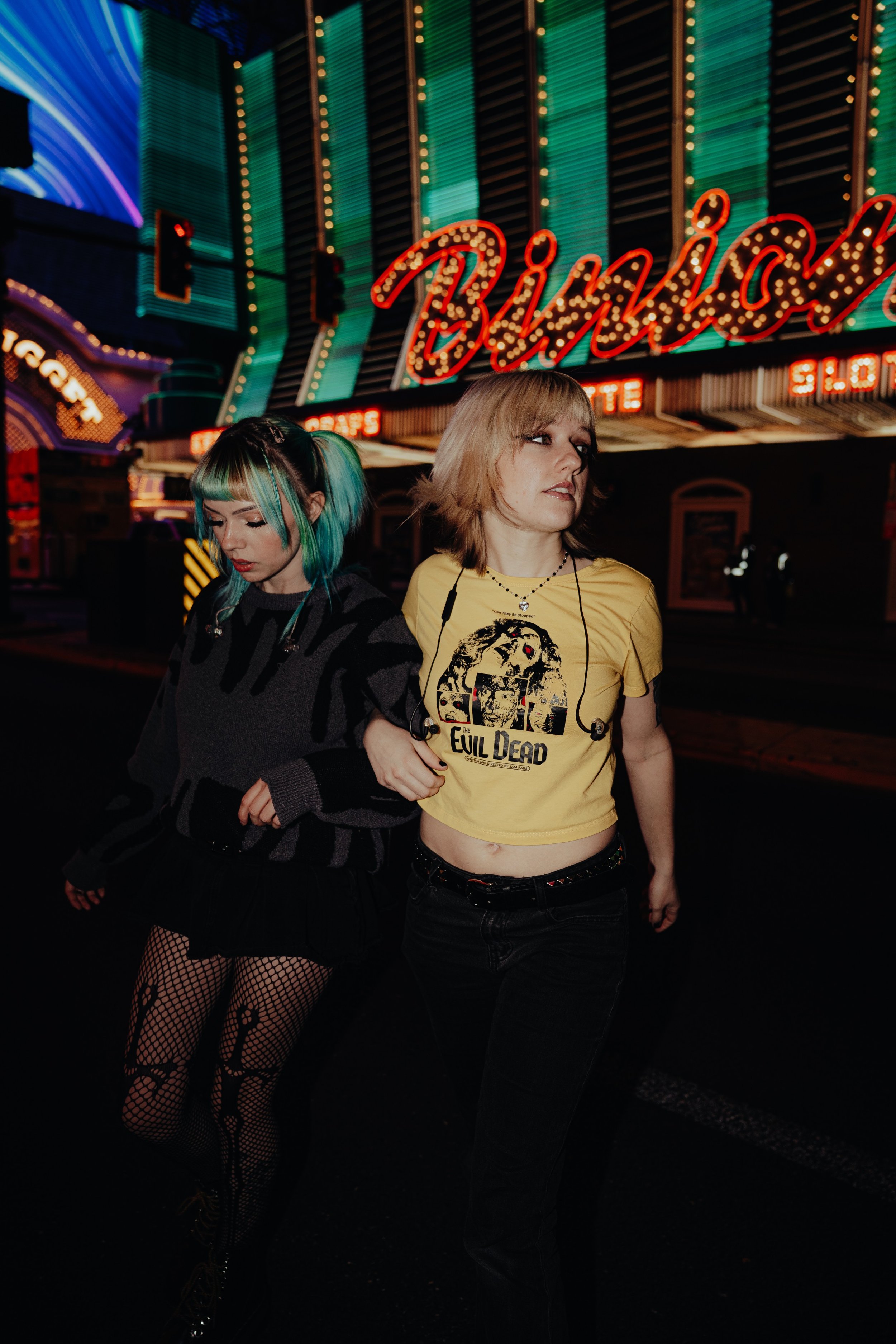 Two young women standing on a city street at night with bright city lights behind them. One has short teal and black hair, wearing fishnet stockings and a dark sweater. The other has short blond hair, wearing a yellow T-shirt with a graphic and black