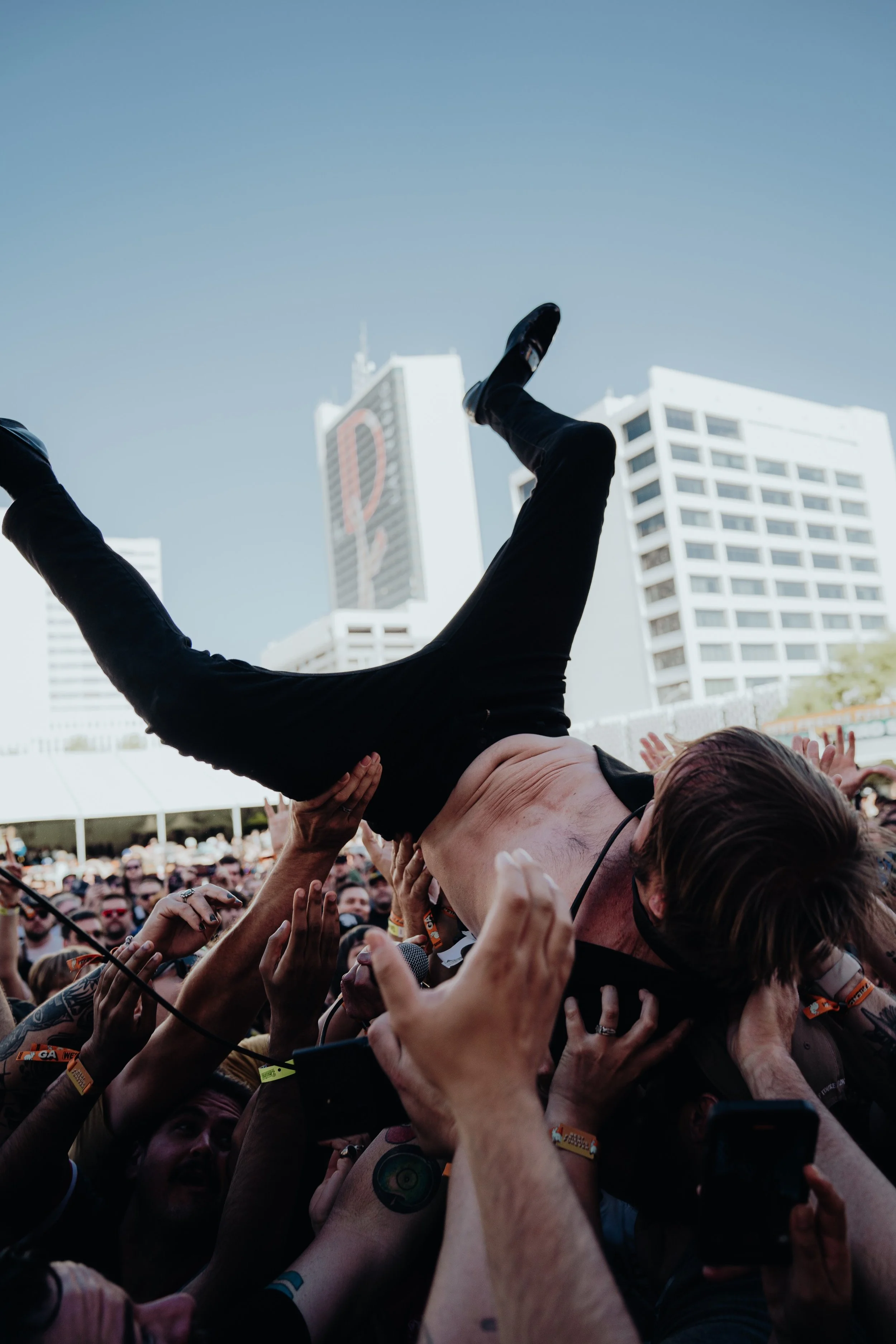 People at an outdoor concert forming a crowd, with one person crowd surfing above the audience against a backdrop of tall buildings and a clear sky.