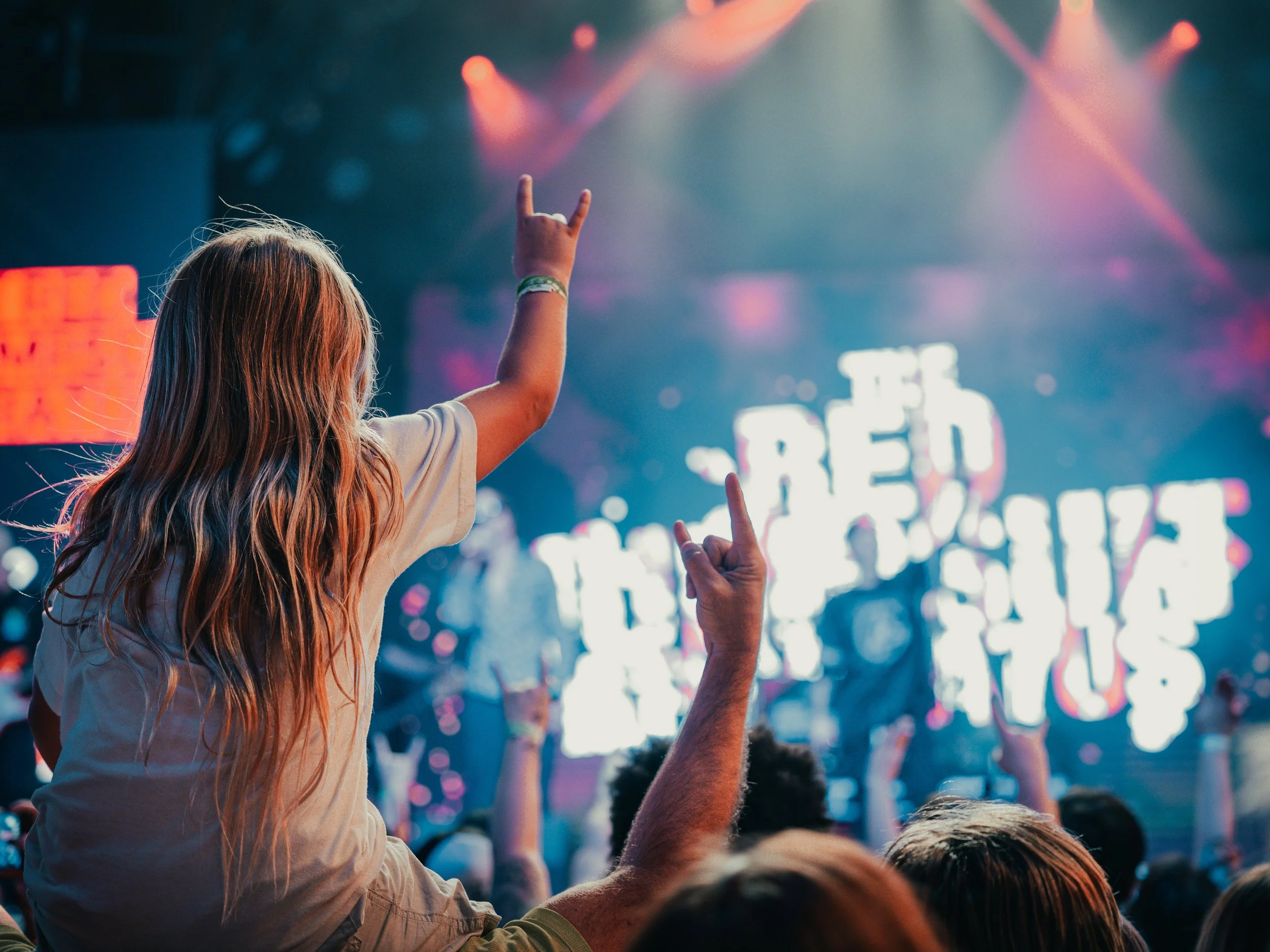 A person with long hair making a rock and roll hand gesture at a concert or event with colorful stage lights and a crowd in the background.