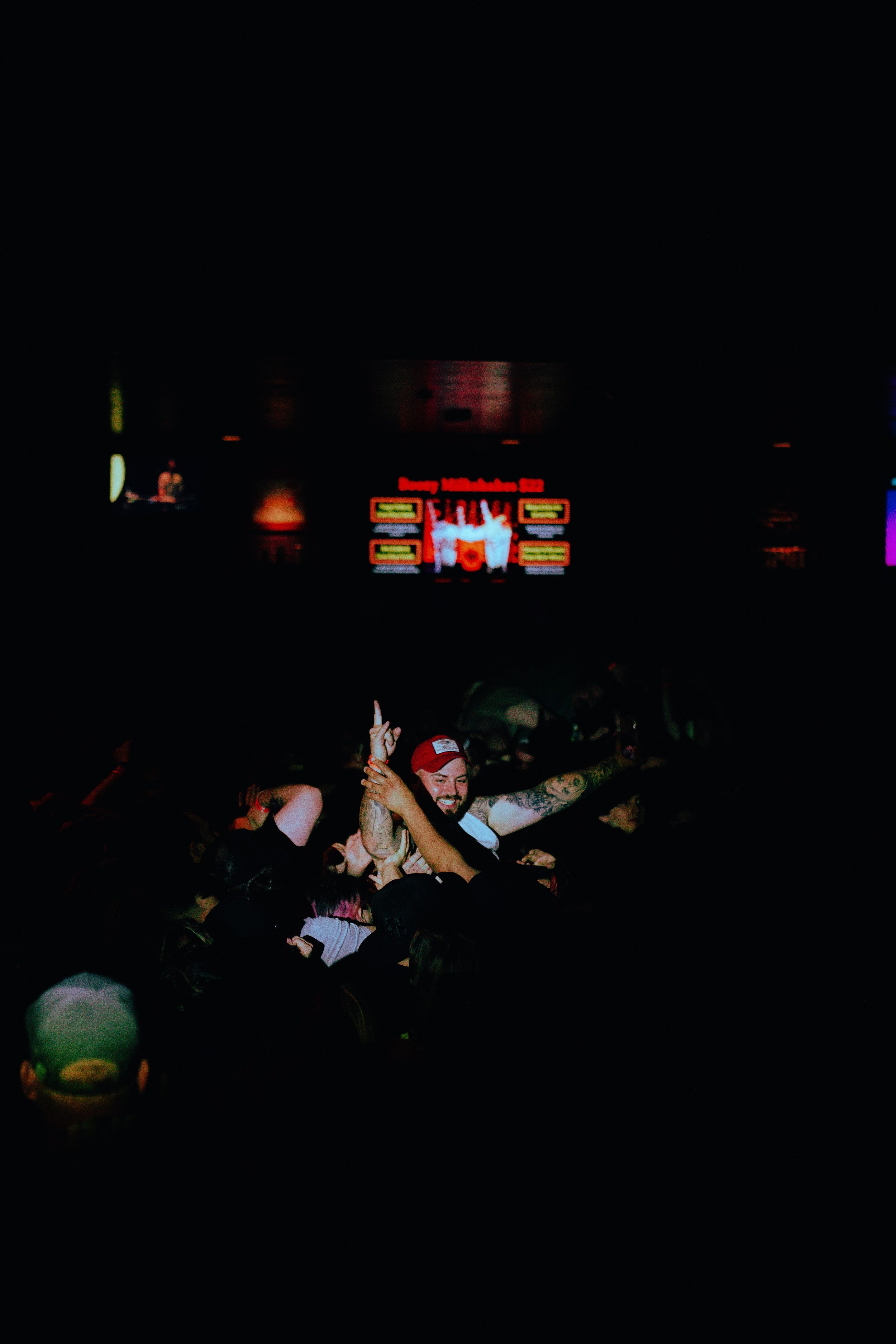 Crowd of people celebrating at a concert or live event with hands raised, some holding drinks, in front of a large screen displaying colorful graphics and text.