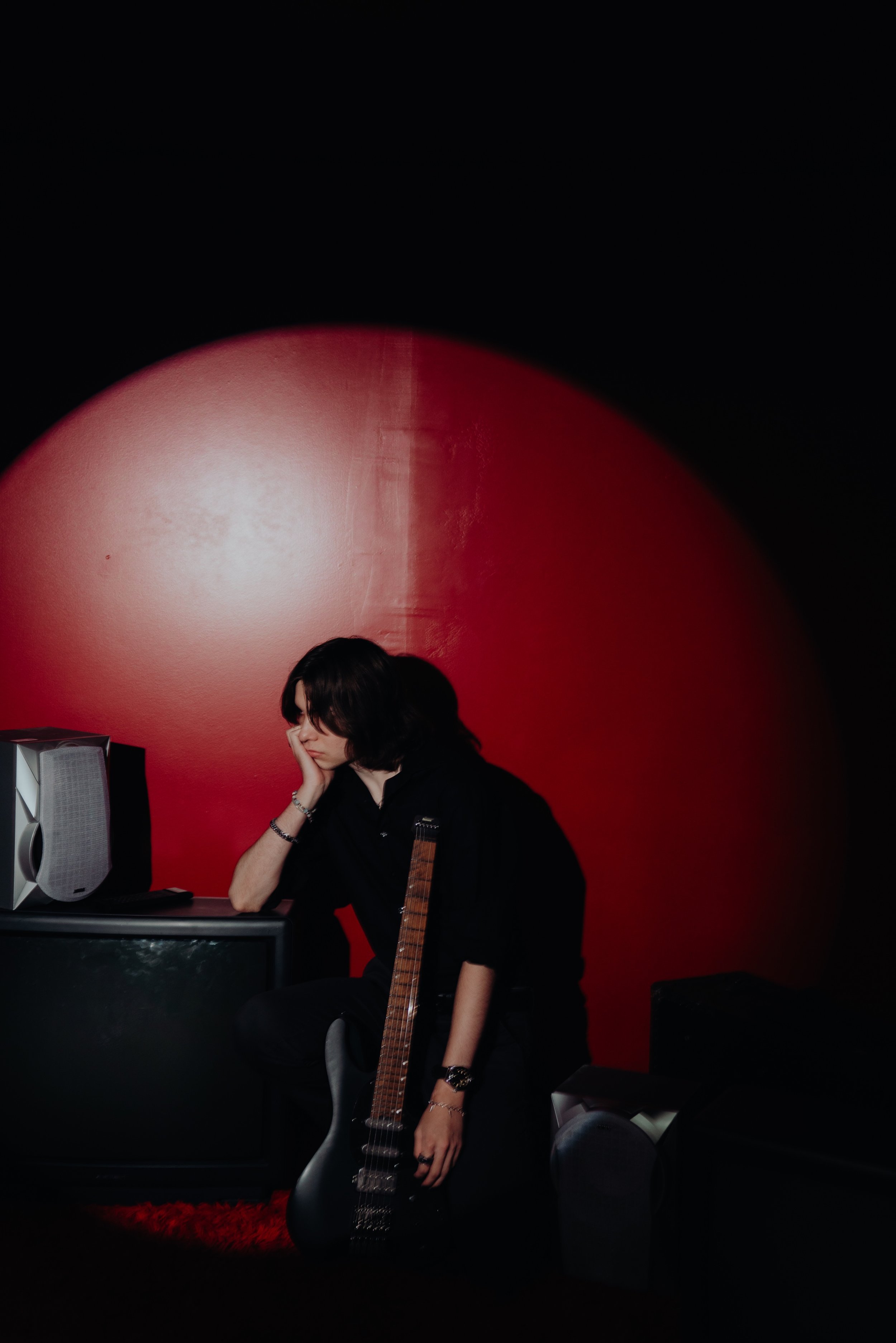 A young woman with dark hair sitting at a desk with a computer, resting her head on her hand, in front of a red wall, with an electric guitar standing beside her.
