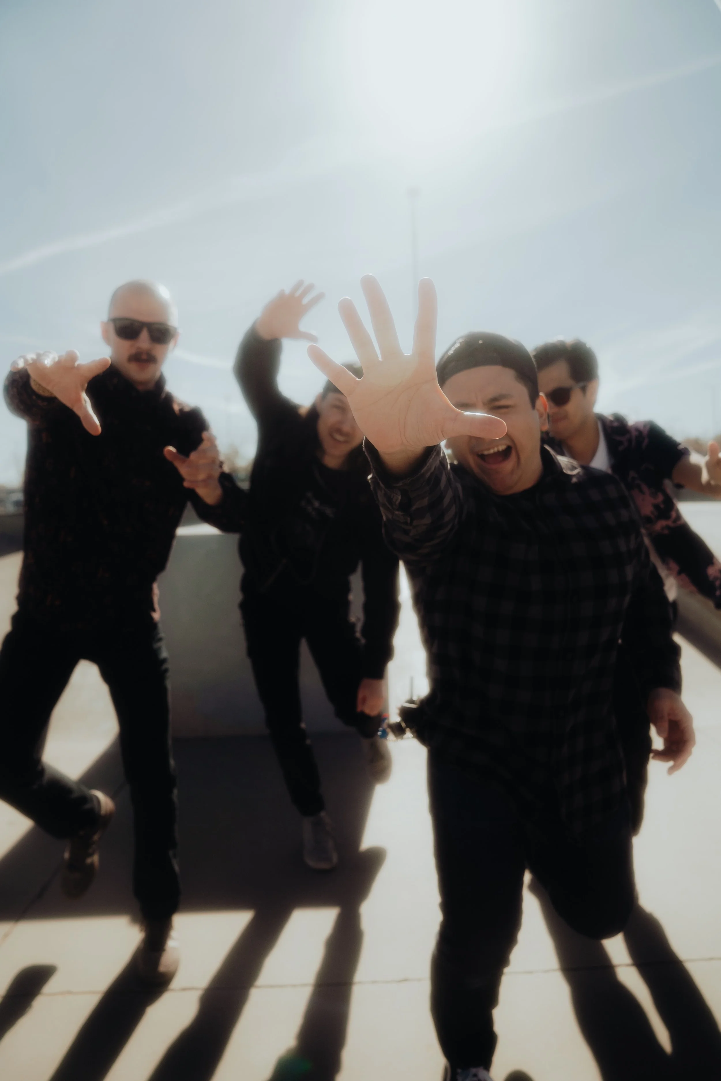 Six people are standing outside on a sunny day, posing for a photo with their hands reaching towards the camera, casting shadows on the ground.