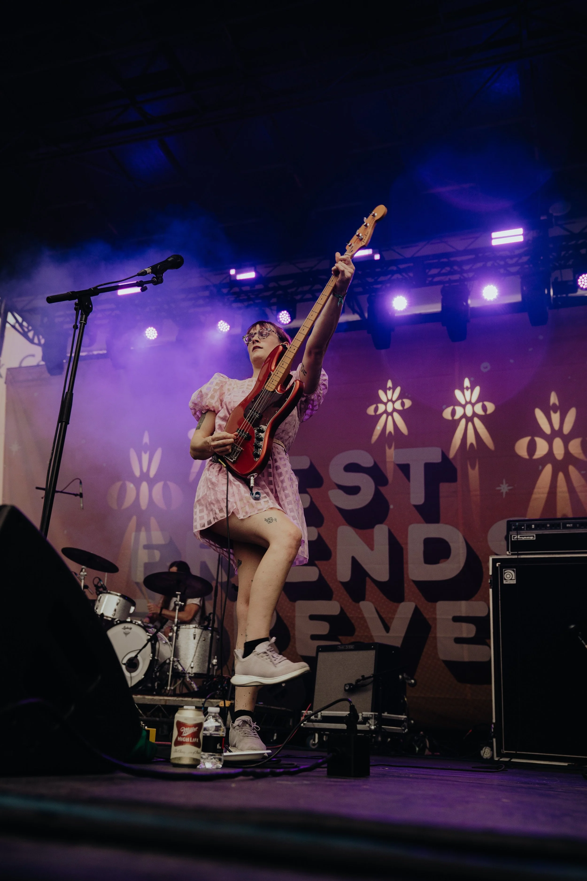 A female musician with glasses, wearing a pink dress, performing on stage with a bass guitar. She stands on one leg, with her right leg raised and positioned on a monitor, near a microphone stand. The background features stage lights and a banner wit