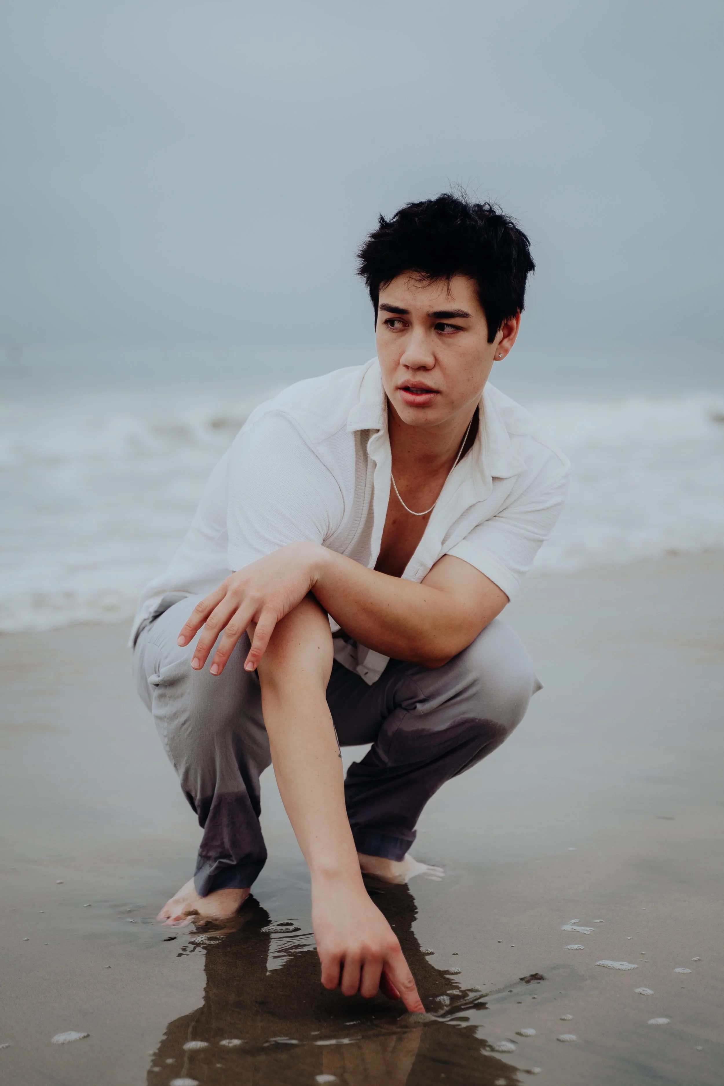 A young man with dark hair and a white shirt crouches on a beach, touching the wet sand with his finger. The sky is overcast, and the ocean waves are in the background.