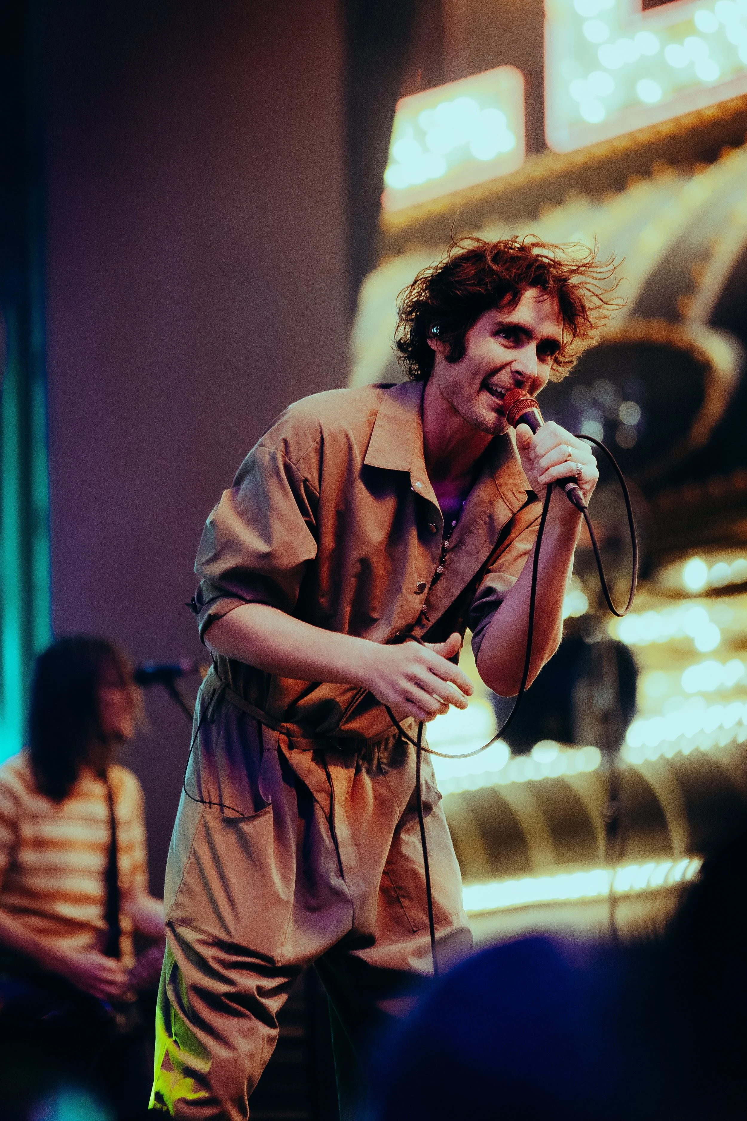 A man with curly hair holding a microphone and singing or speaking in a dimly lit neon-lit setting, with a woman in the background.