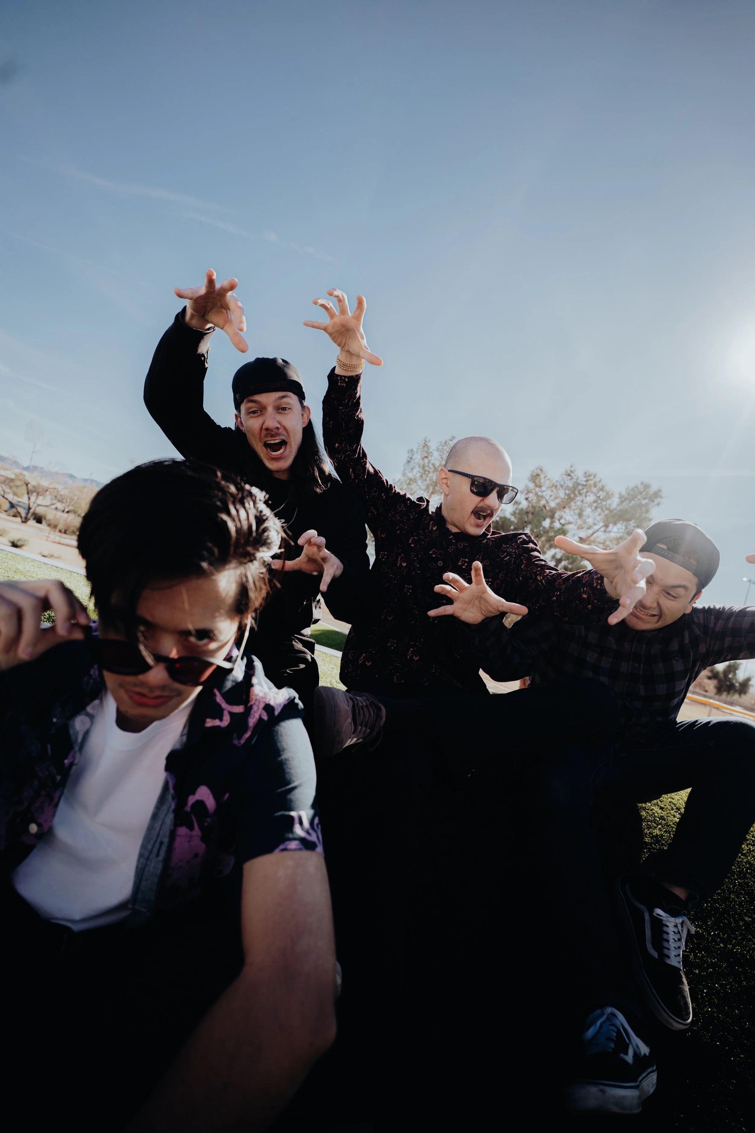 A group of people outdoors, some with their hands raised, making expressive gestures, and smiling. The photo is taken from a low angle with a clear blue sky in the background, suggesting a sunny day.