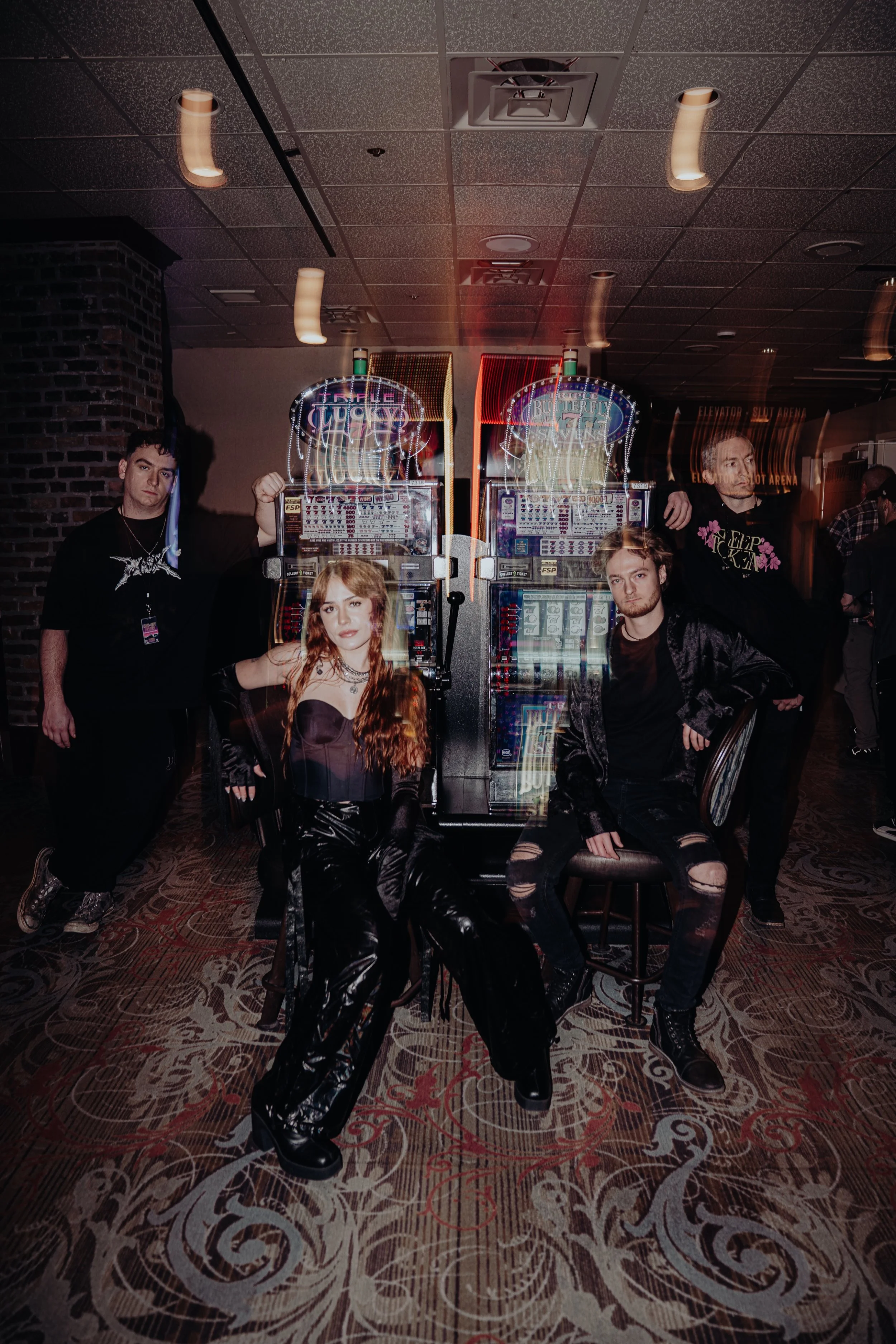 Five young adults posing in front of slot machines in a casino. Three men and two women, dressed in dark clothing, with the women seated and the men standing behind them.