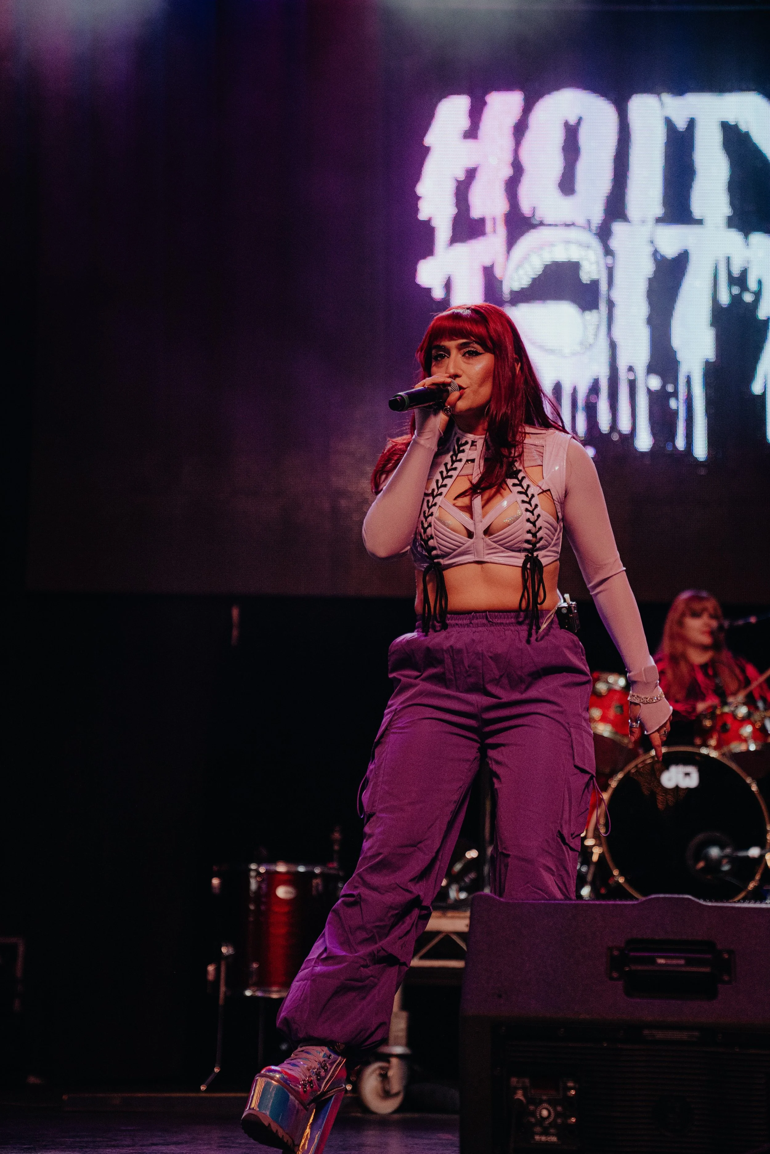 A performer with long red hair singing into a microphone on stage with a neon sign in the background.
