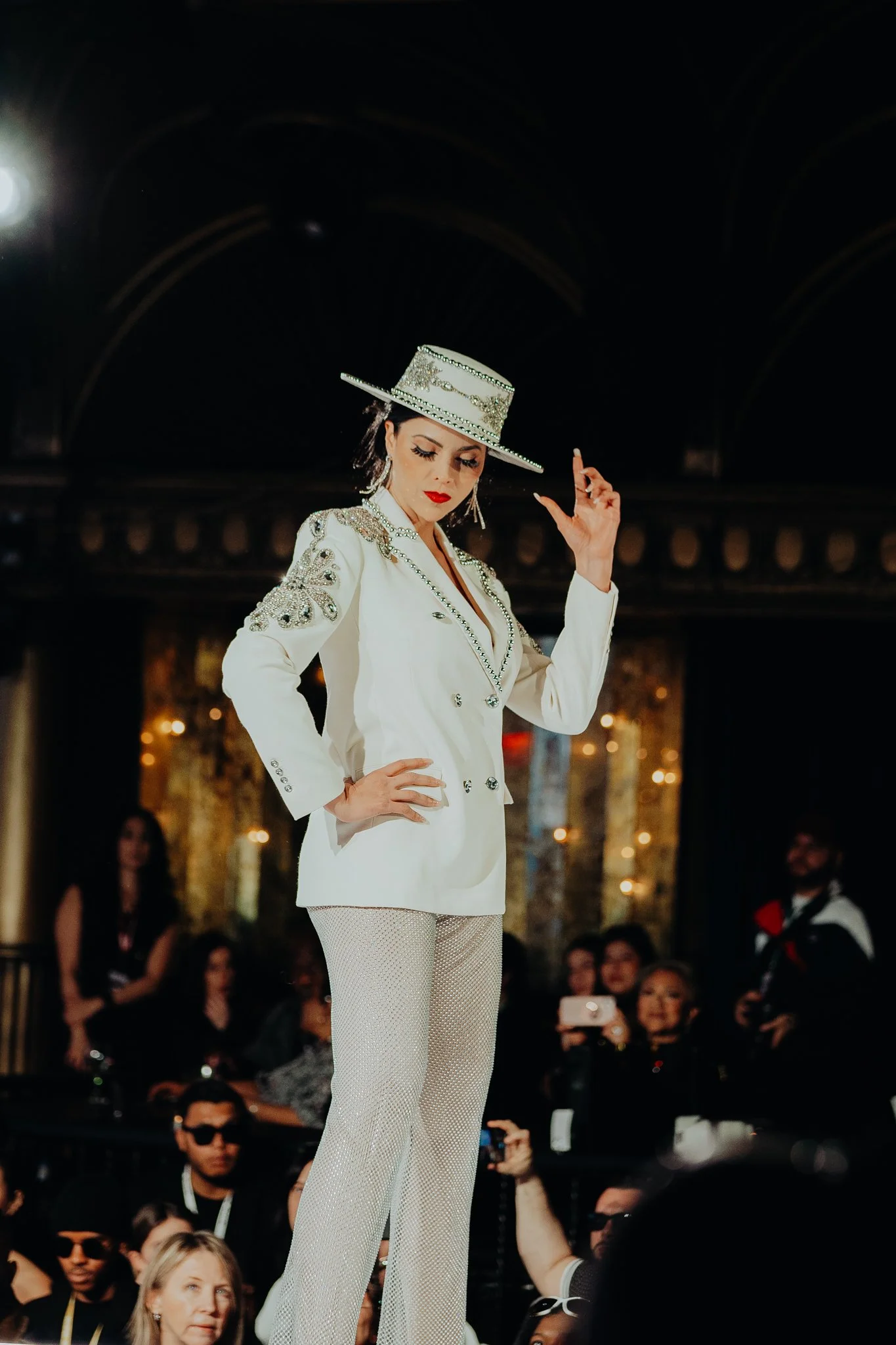 A woman walking runway at a fashion show, wearing a white suit with intricate embellishments and a matching wide-brimmed hat with decorative accents. She has dark hair, bright red lipstick, and is striking a confident pose with her head slightly tilt