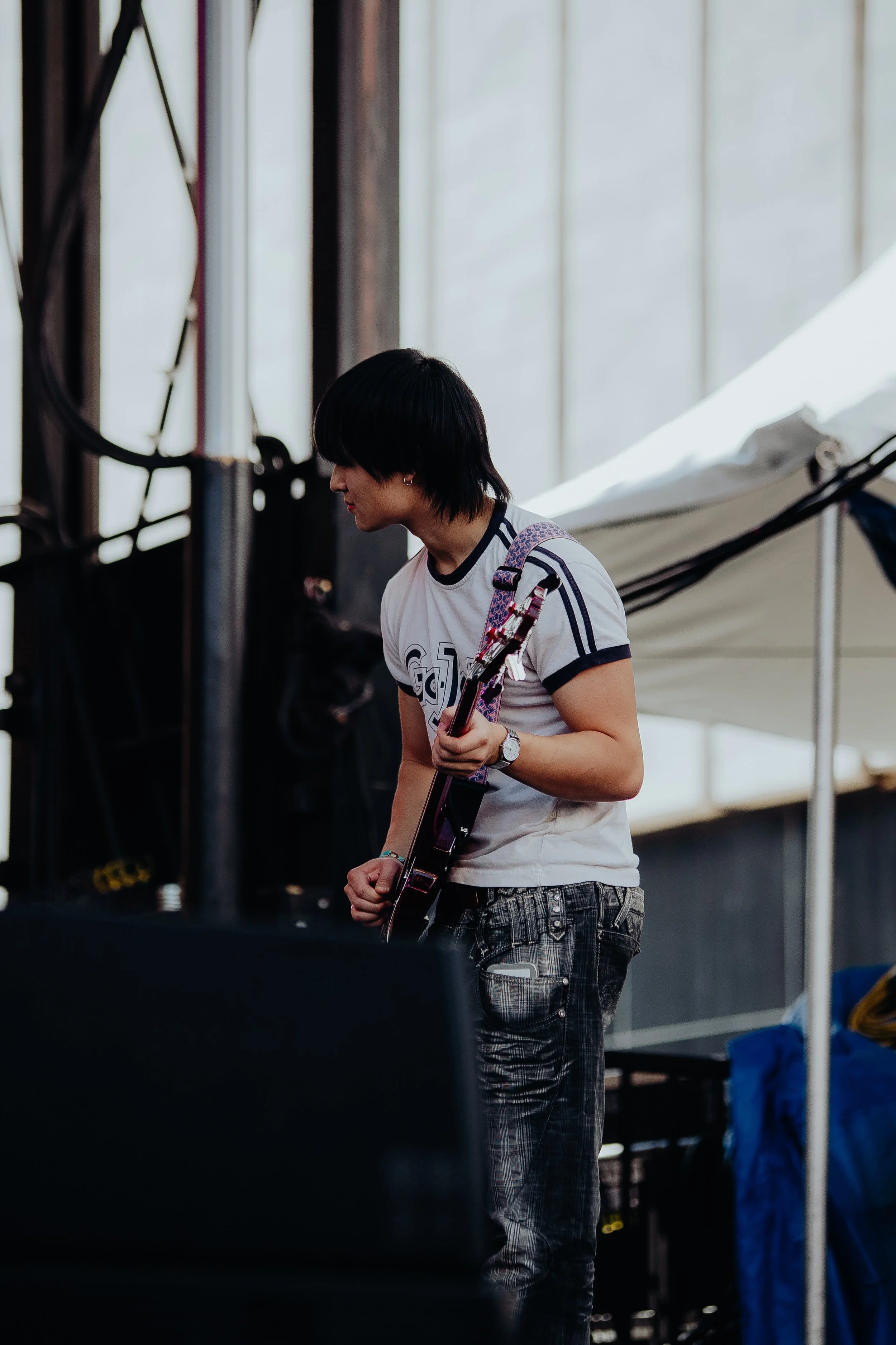 A woman with short dark hair wearing a white t-shirt with black trim and patterned pants, holding a guitar, standing near a stage setup in an indoor setting.