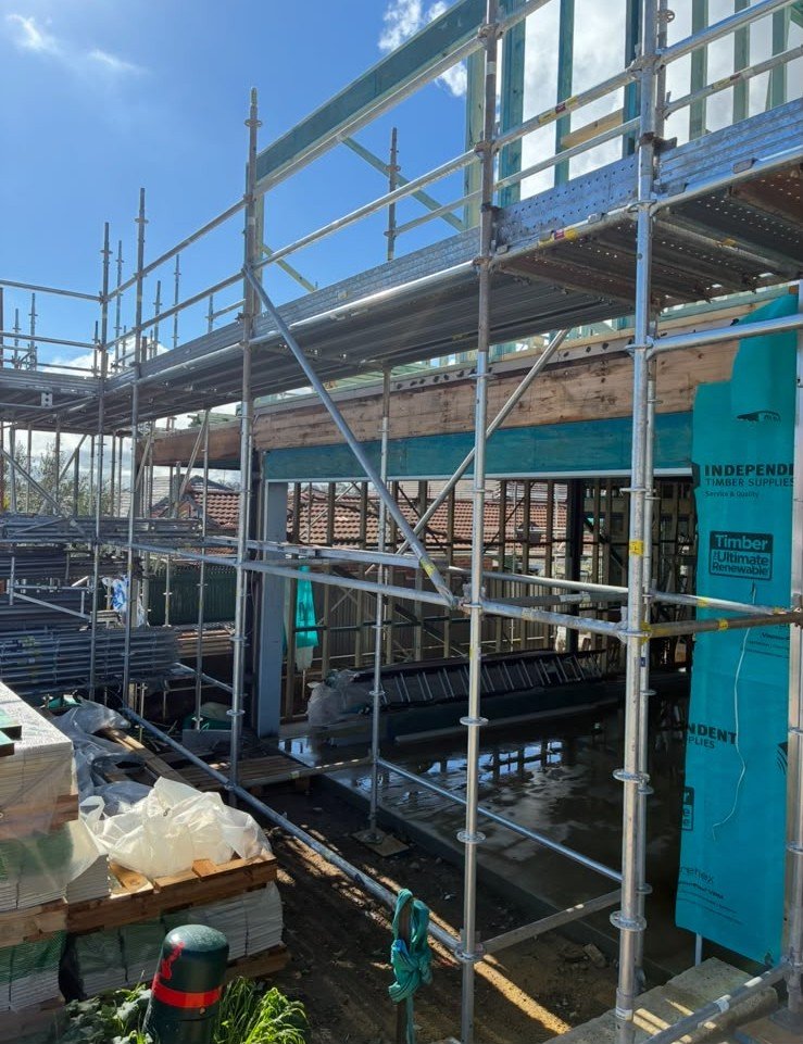 Construction site with scaffolding around a building frame, with building materials and tools in the foreground, and a clear blue sky overhead.