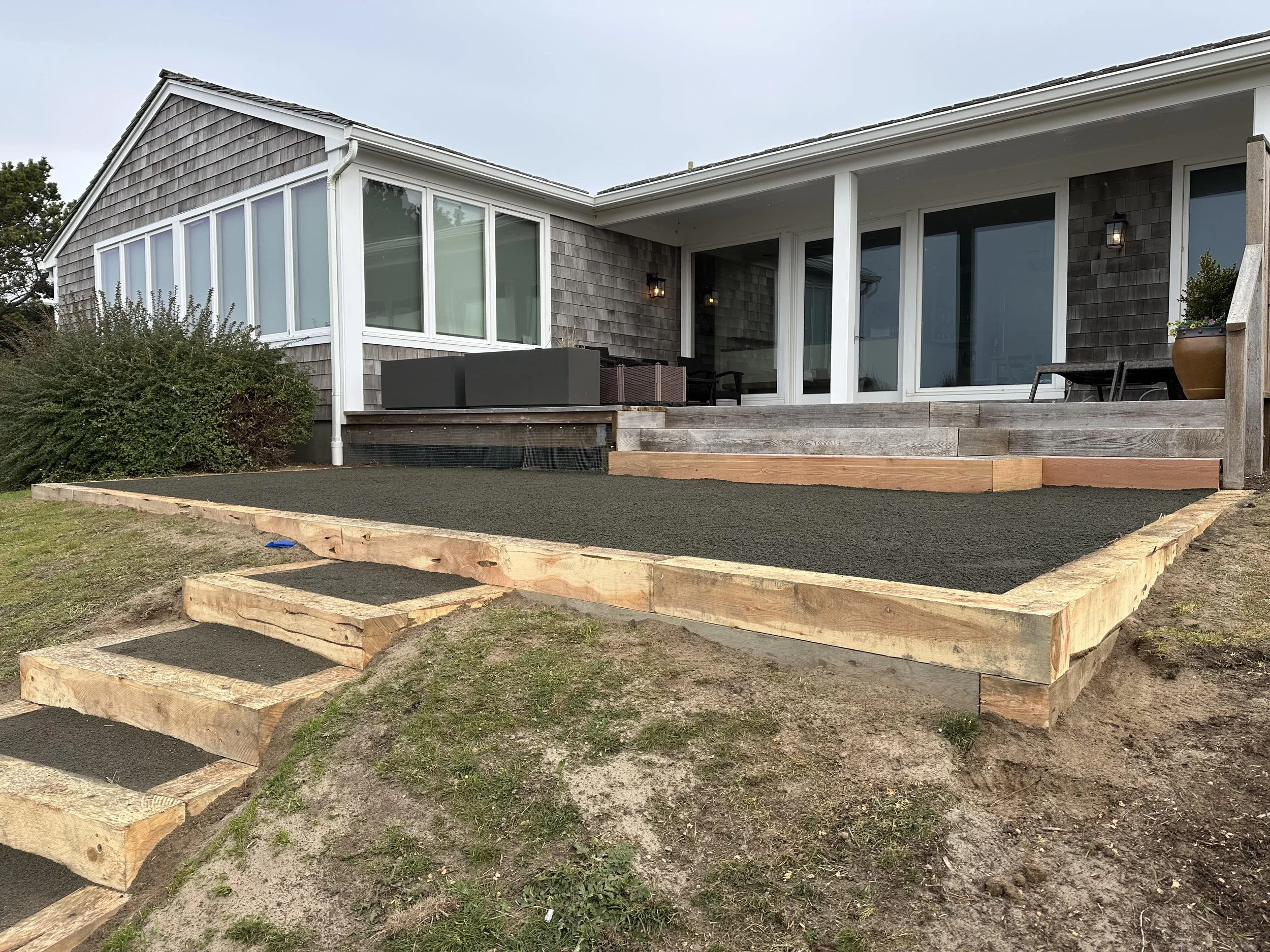 A house with wooden shingles and large windows has a newly constructed wooden deck and stairs leading up to the patio. There are outdoor lights on the house, and some bushes and grass in the yard.