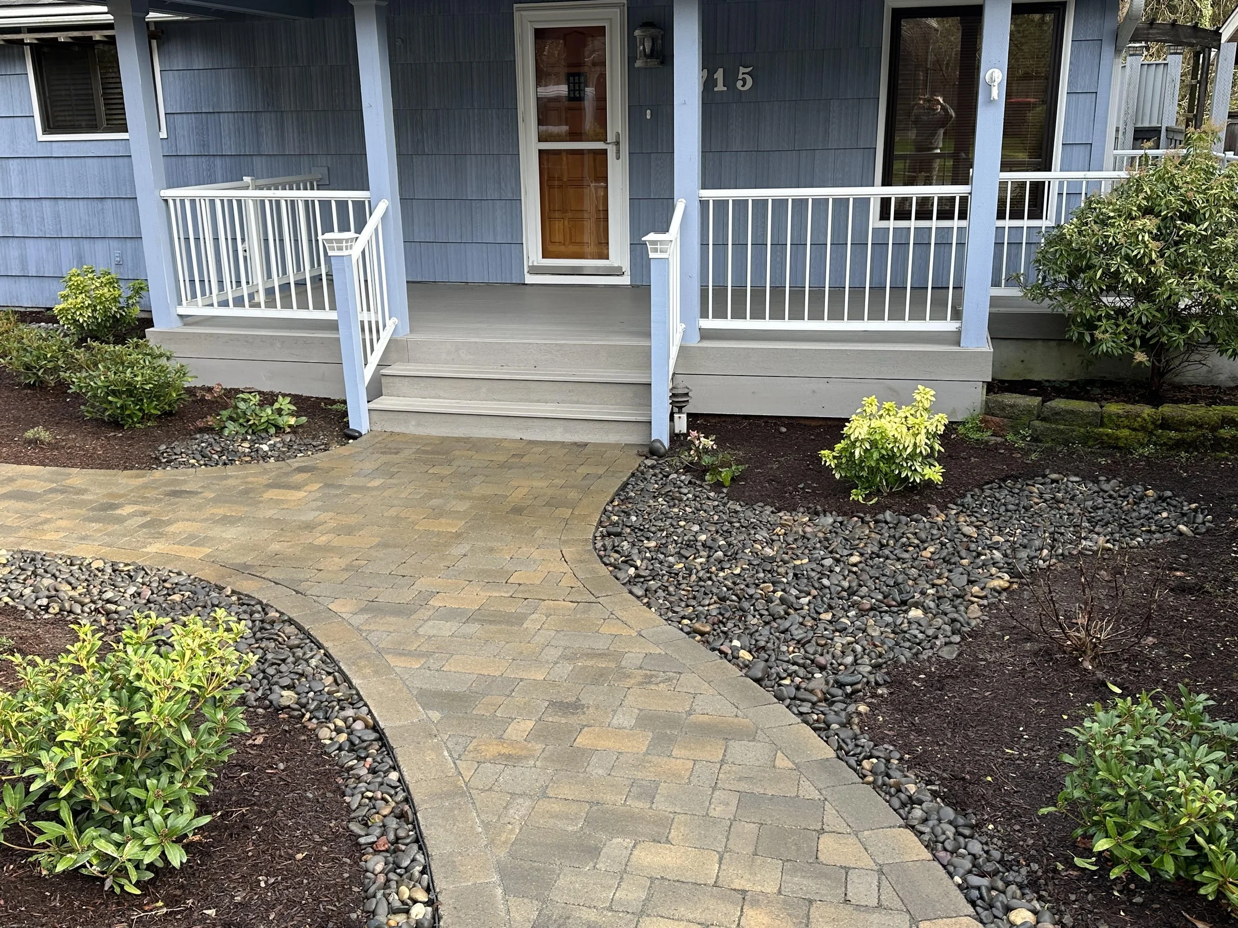 Front porch of a blue house with stairs, white railing, and a brown door surrounded by landscaped plants and rocks.