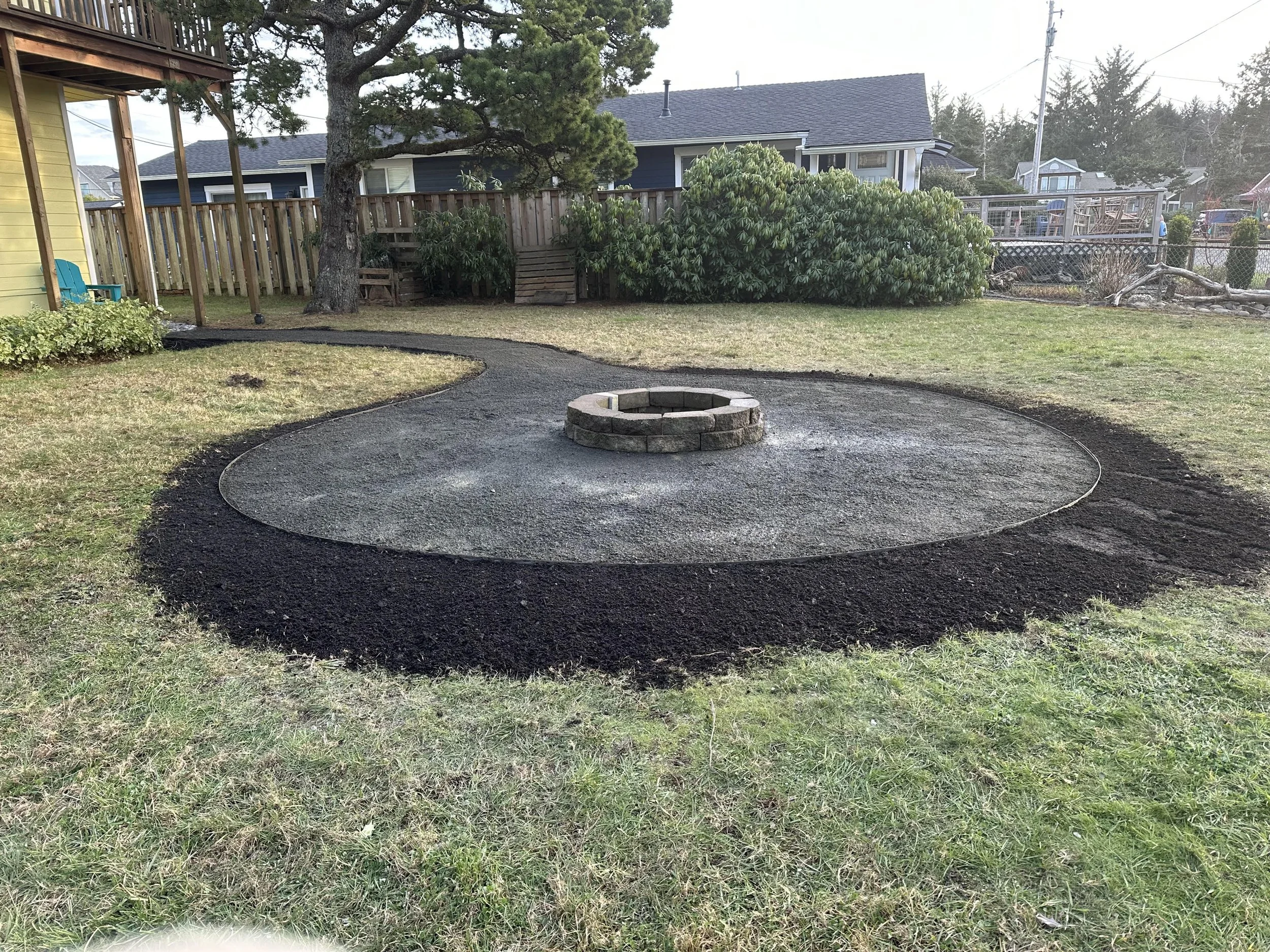 Backyard with a fire pit, patchy grass, a tall tree, a wooden fence, and neighboring houses with decks and trees.