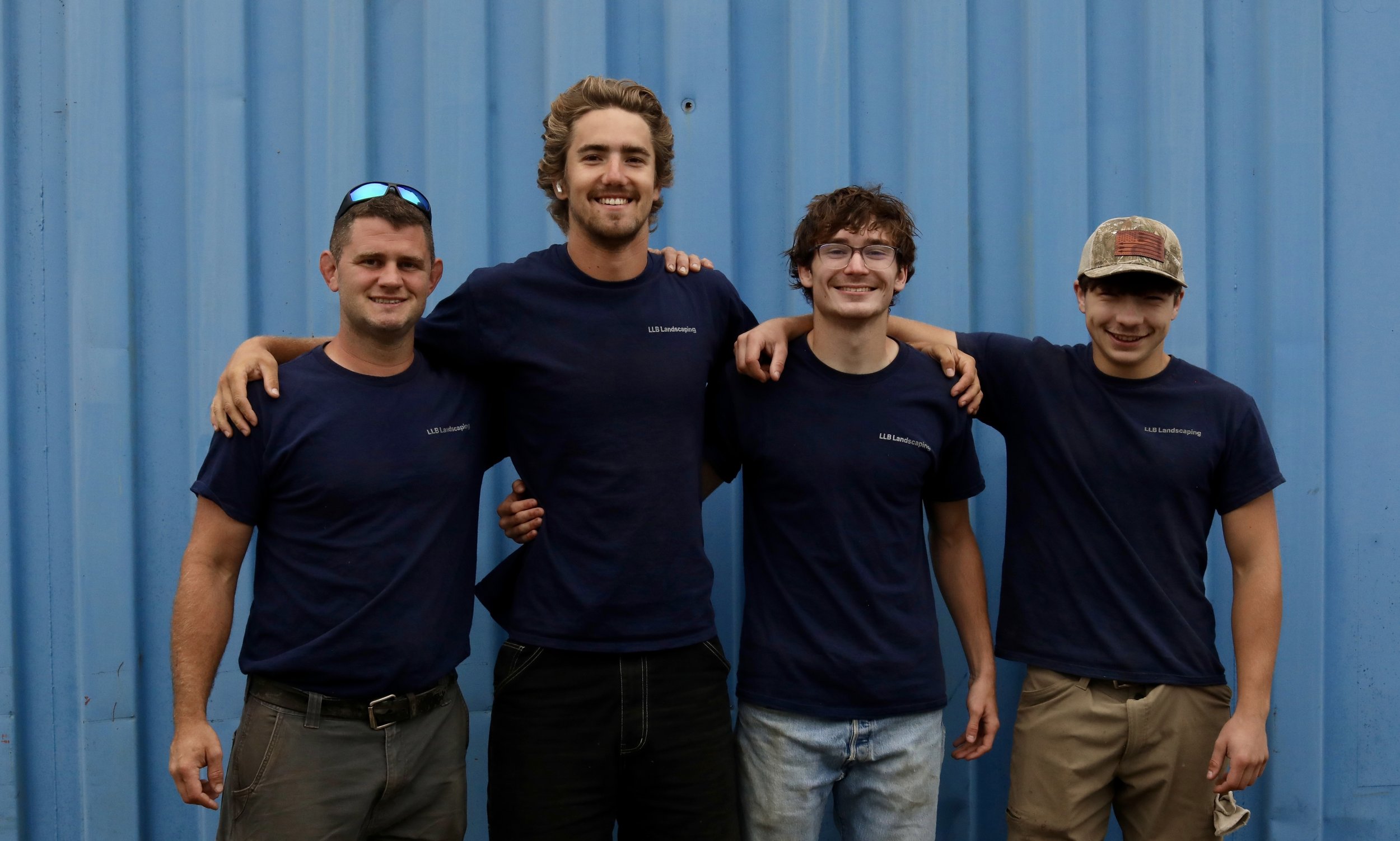 Four men standing close together with their arms around each other against a blue corrugated metal wall, smiling at the camera. All are wearing navy blue shirts with the logo 'LLB Landscaping'.