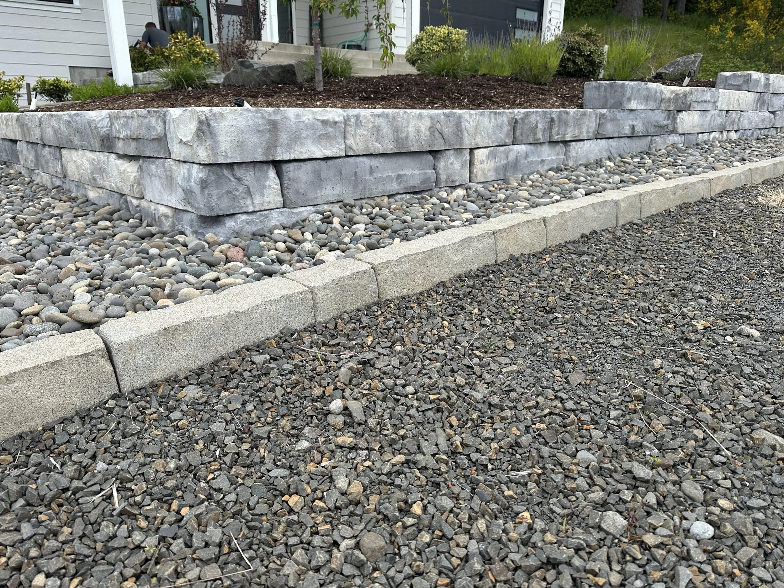 A stone retaining wall with a beds of small multicolored gravel and larger stones in front of a landscaped yard with shrubs and flowers, and a person working near the house in the background.