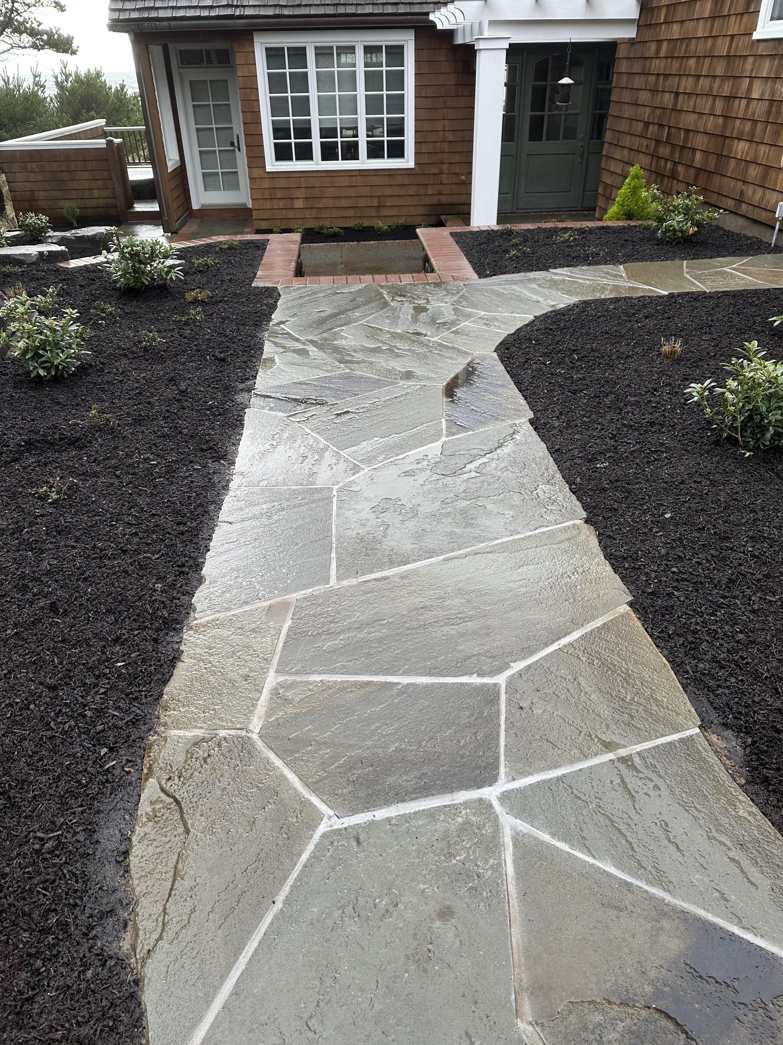 A backyard with a stone path, landscaped garden beds, and a house with wooden siding and multiple windows.
