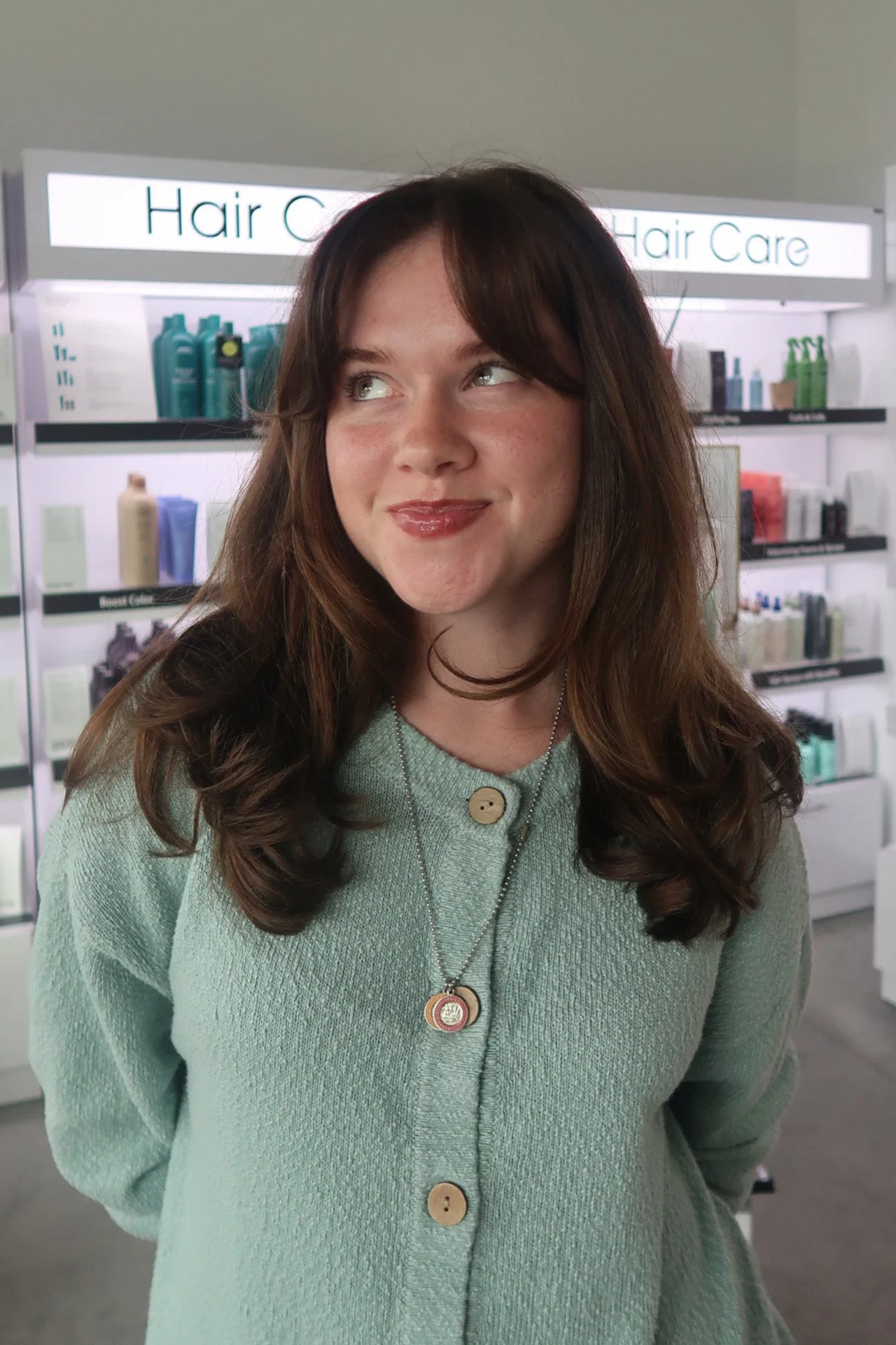 A woman with long brown hair and a light green sweater standing in front of a hair care product display in a store.