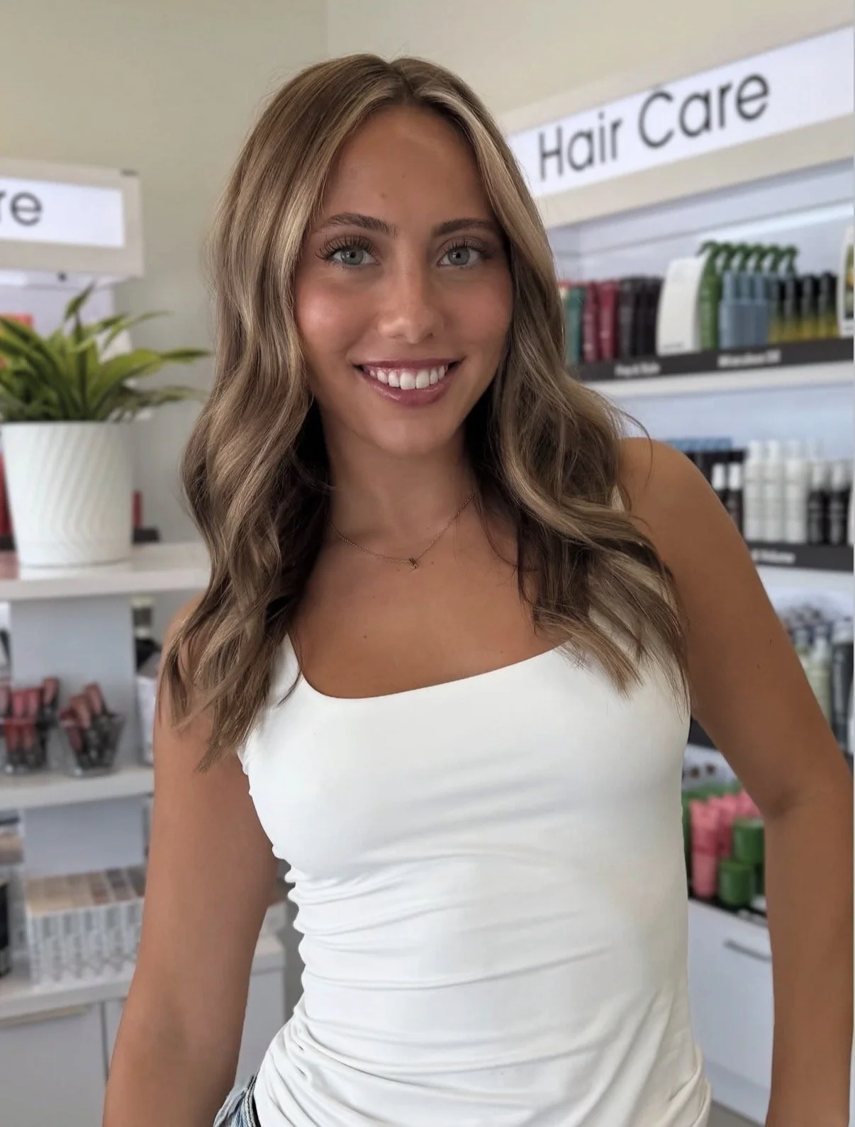 A smiling young woman with long wavy blonde hair and blue eyes, wearing a white sleeveless top and a delicate necklace, standing in a store aisle with hair care products and a potted plant in the background.