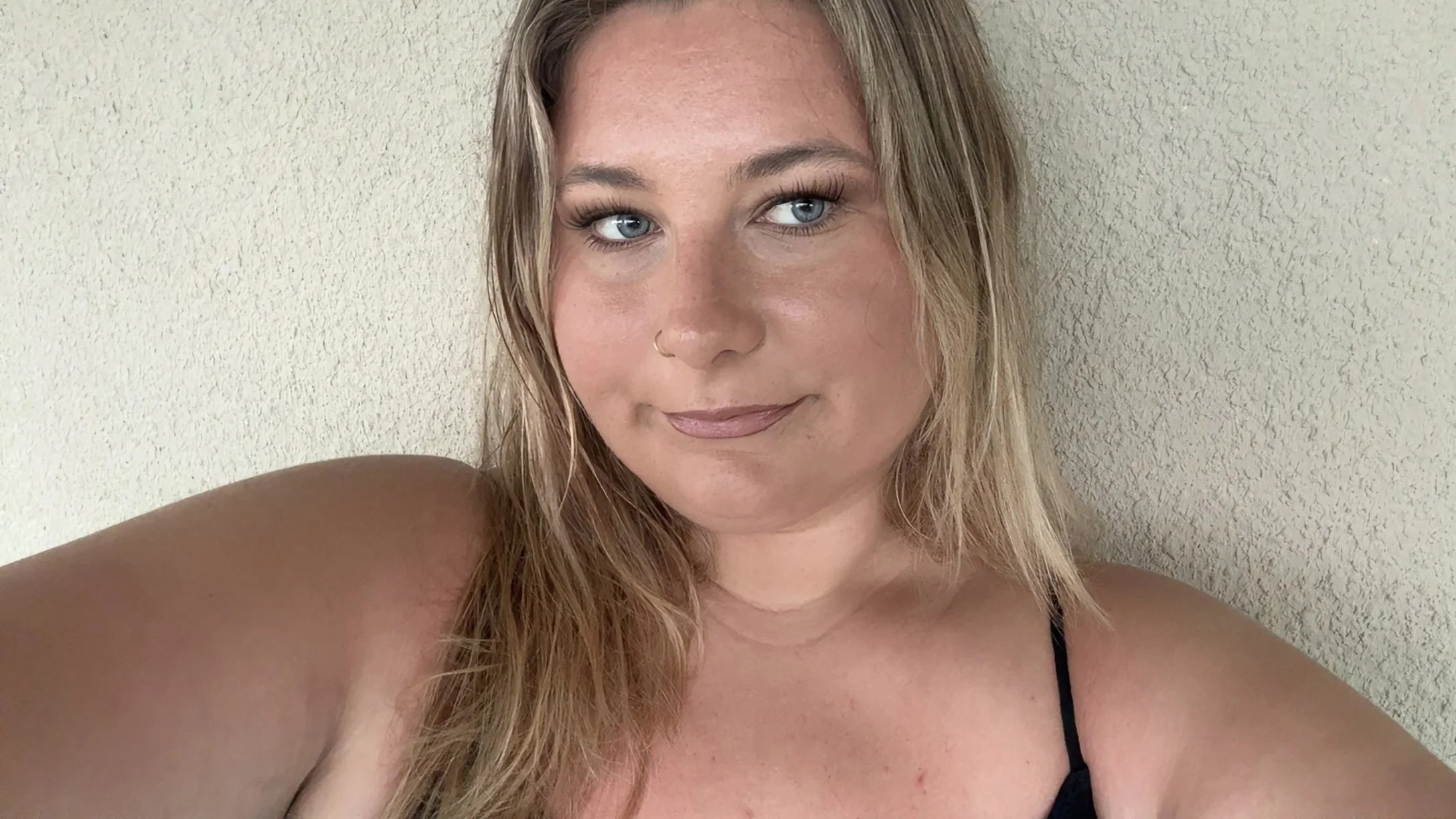 A woman with blonde hair and blue eyes taking a selfie against a beige wall, wearing a black strap top.