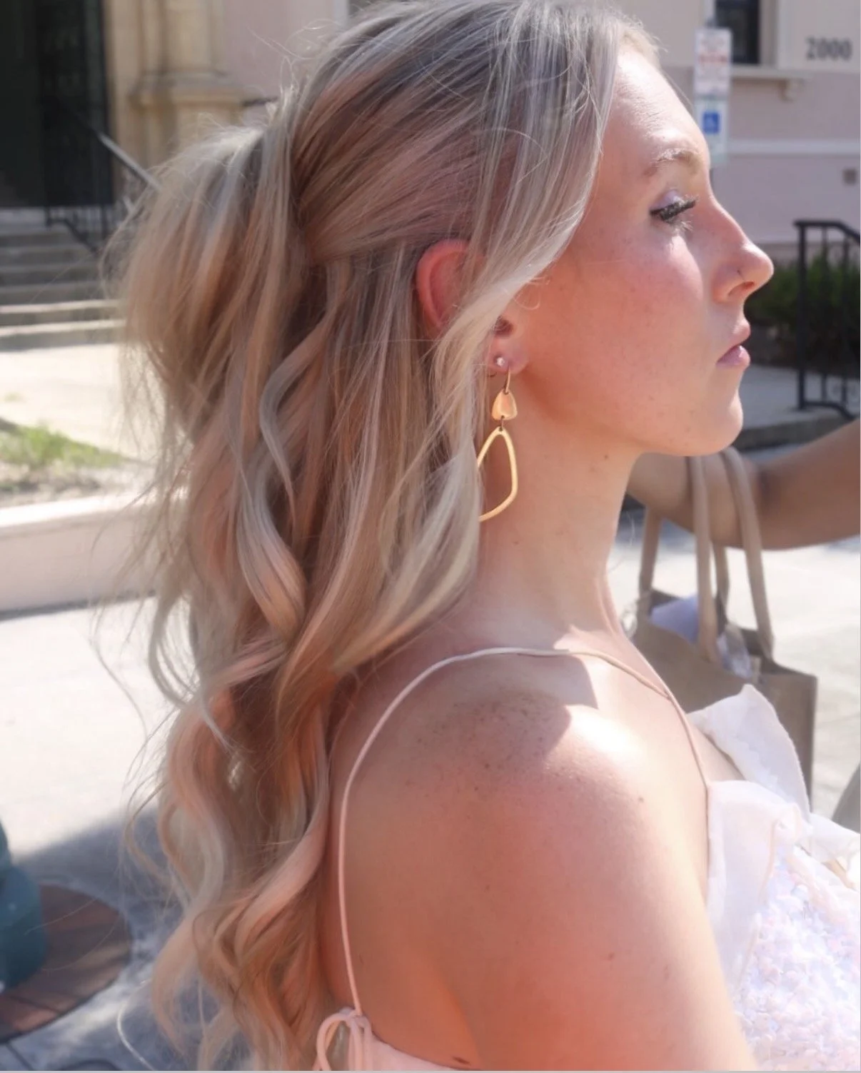 A woman with long, wavy blonde hair, wearing gold teardrop-shaped earrings, in profile, outdoors in sunlight, with stairs and a building in the background.