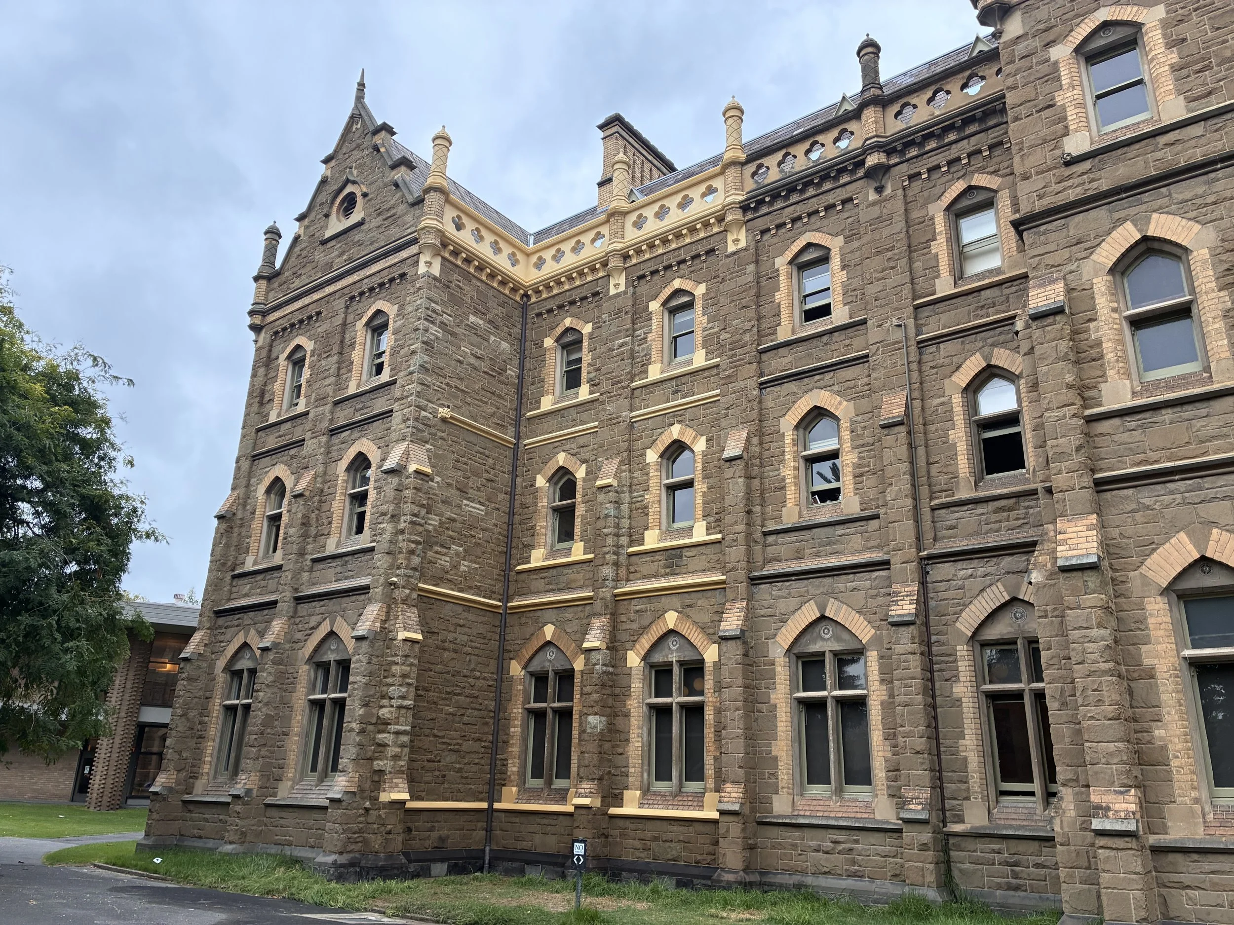 A historic stone building with pointed towers and arched windows, featuring decorative brickwork and a narrow cream-colored balcony along the roofline, set against a cloudy sky.