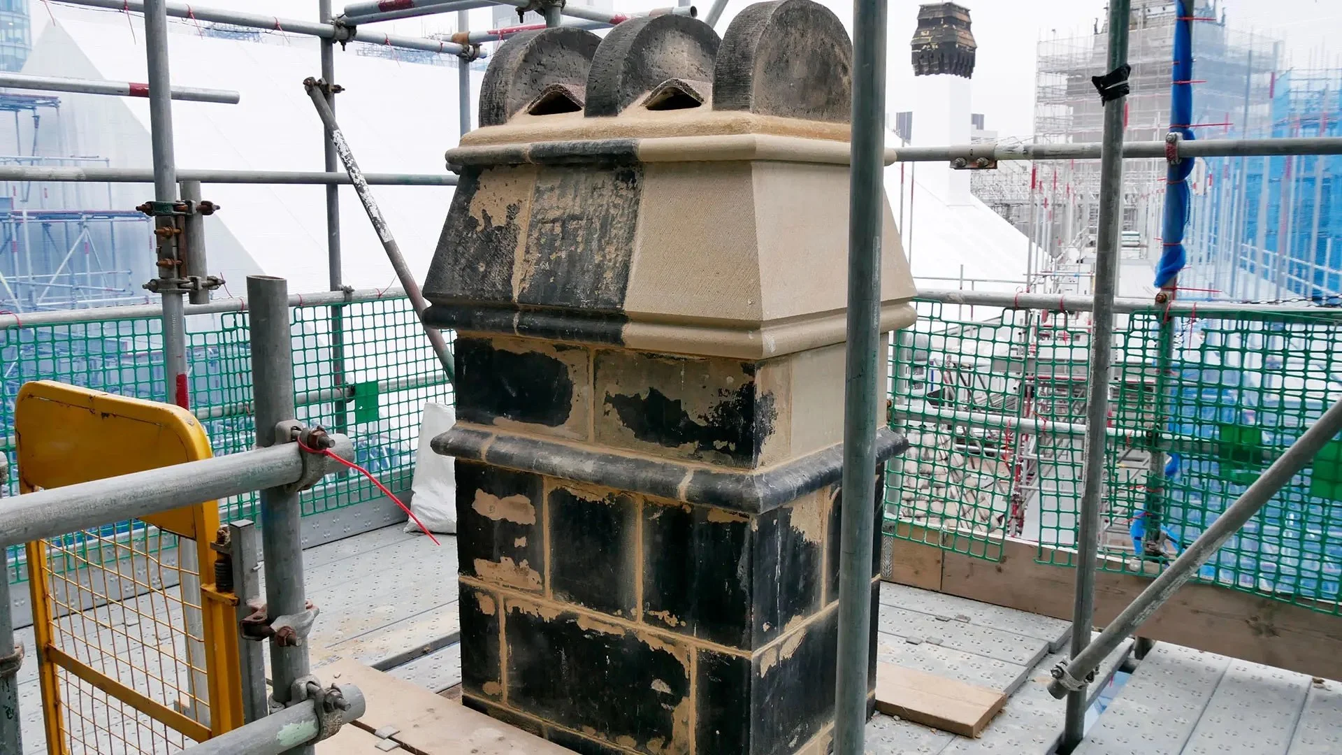 A construction site with scaffolding and a stone chimney. The chimney has black soot stains and new beige stones added to it. Safety barriers and green mesh fencing surround the area.