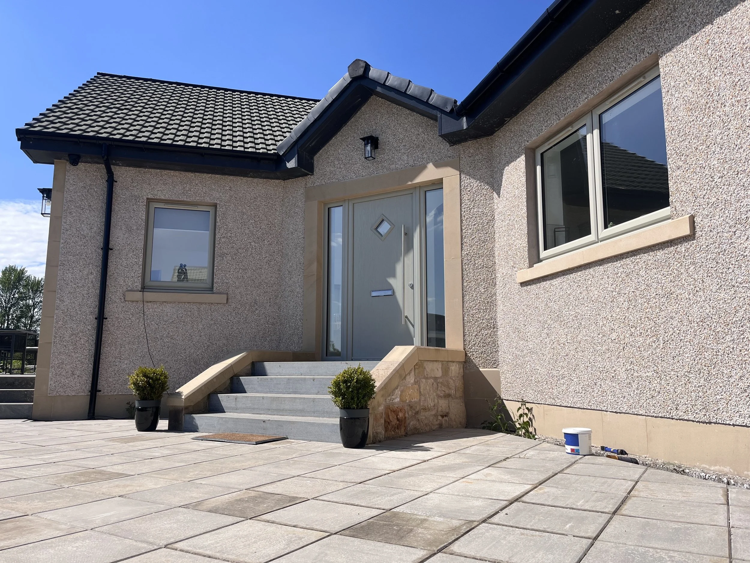 The exterior of a modern house with a tiled roof, large windows, and a beige textured stucco facade. There are steps leading to a front door with a diamond-shaped window, and potted plants on either side of the steps. The front yard has a paved area 
