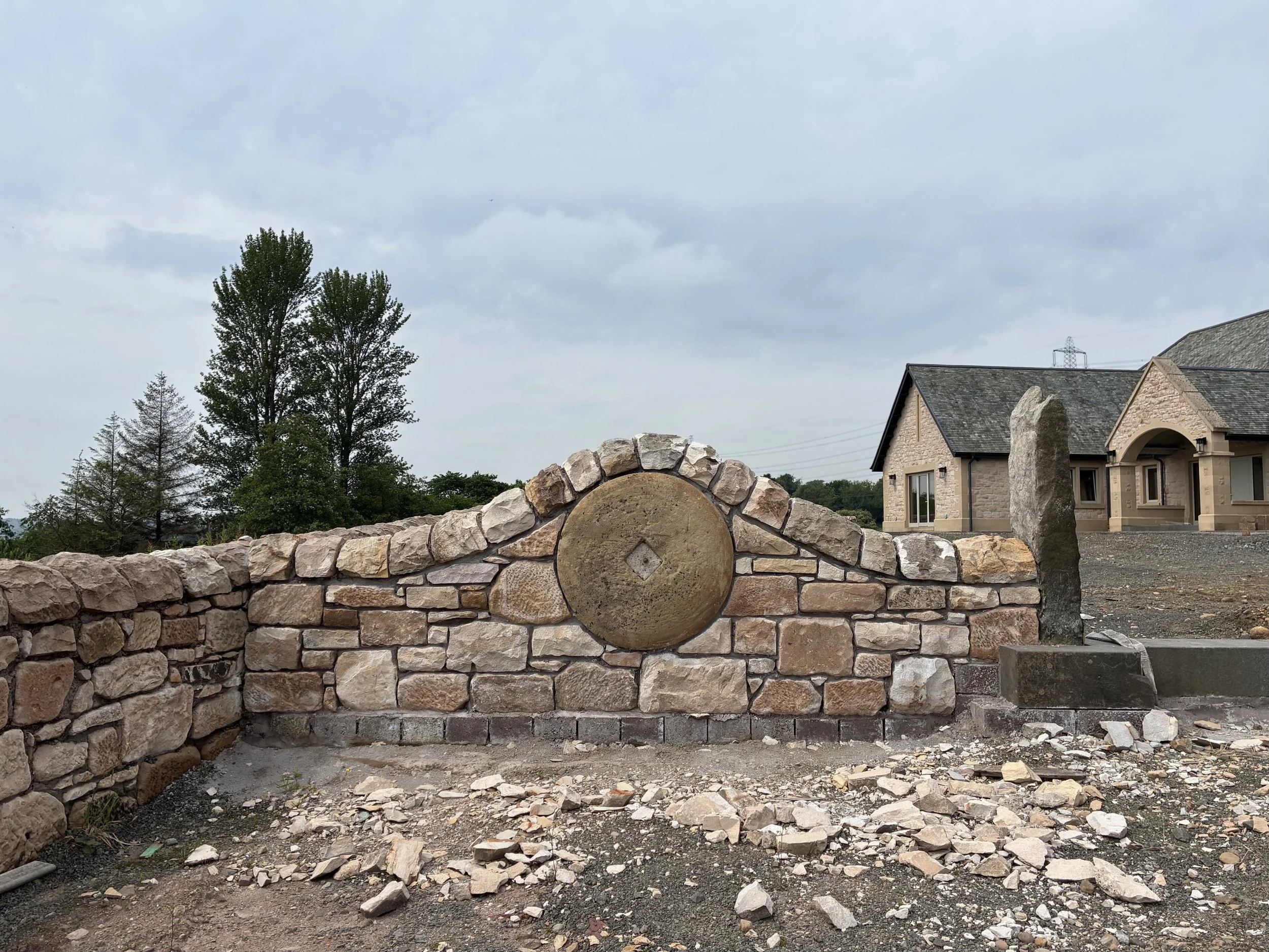 A partially constructed stone wall with scattered loose stones in front, in a residential area with houses, trees, and cloudy sky in the background.