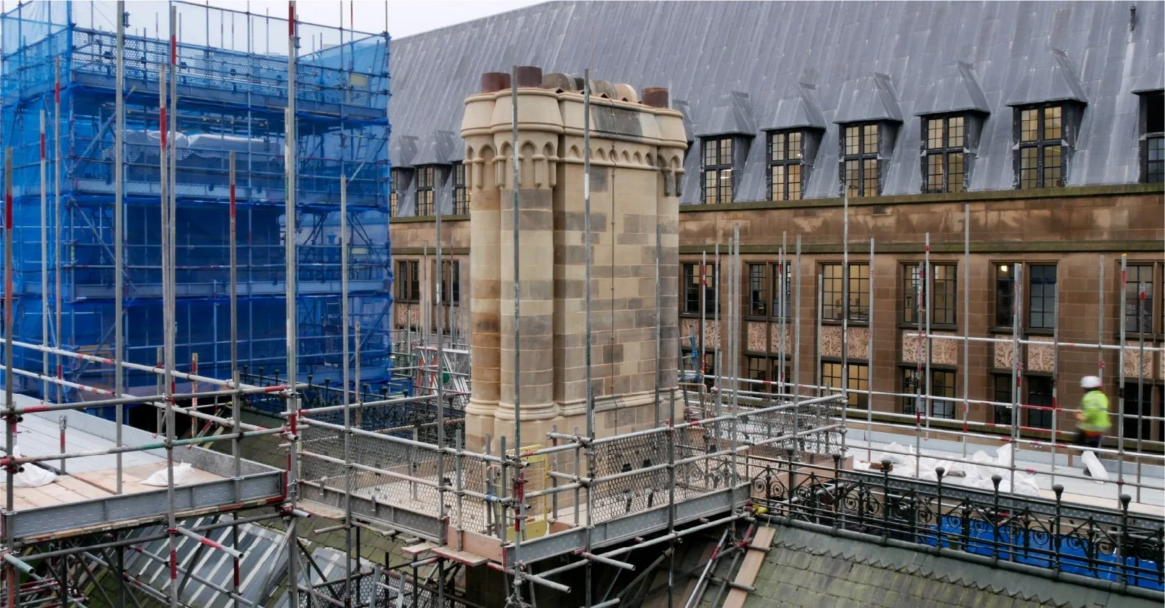 Construction site with scaffolding surrounding a historic building's chimney and roof. A worker in a yellow safety vest and helmet is walking nearby.
