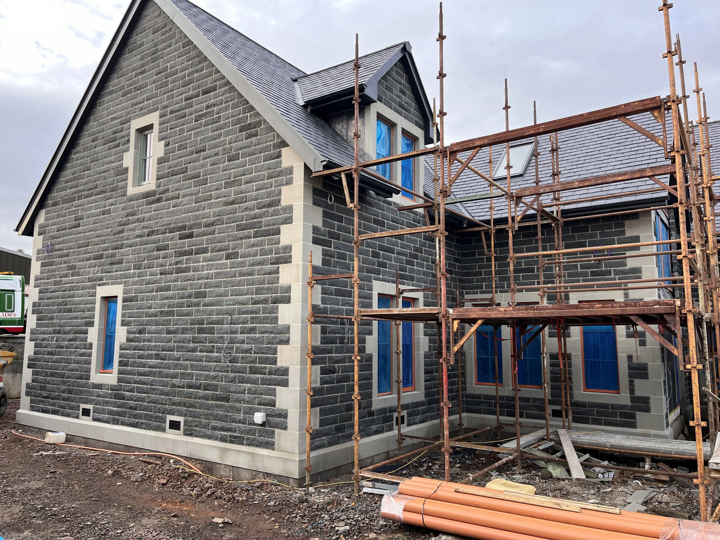 A house under construction with gray brick exterior, scaffolding on the side, and several windows with blue covering. The roof is finished with dark gray shingles.